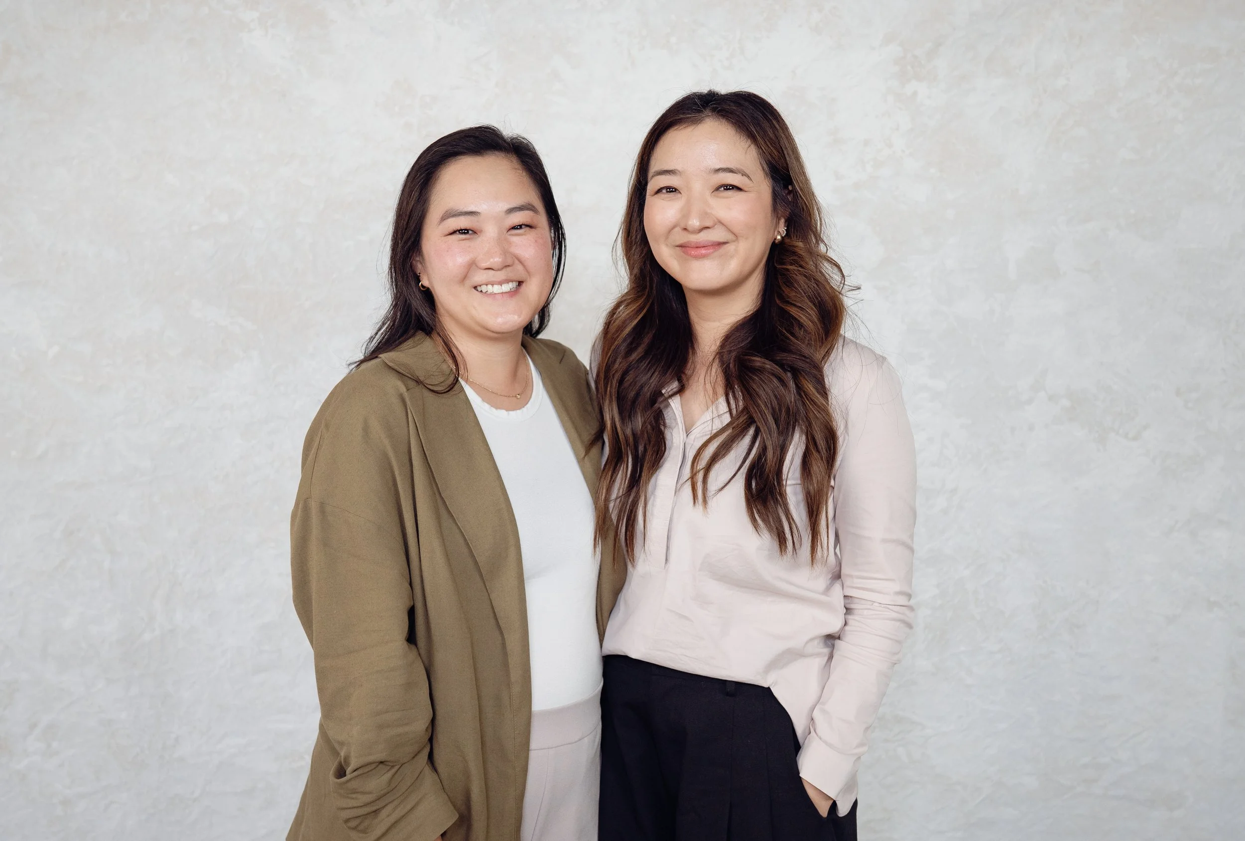 Two women standing close together and smiling in front of a light textured wall.