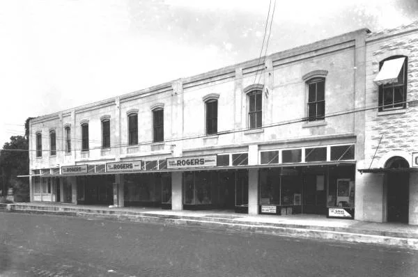 Black and white photo of a two-story historic building with stores on the ground level, including Rogers  store, and apartments or offices on the upper level.