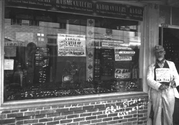 A gentleman standing outside of 115 Main Street with a sign that says 'Champion Whittler of Arkansas Ka-Bar'. Heavy Ka-Bar marketing in the window.