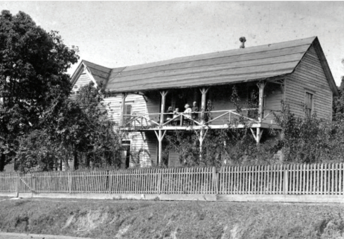 Black and white photo of a two-story wooden house with a porch, surrounded by trees and a wooden fence. People are standing on the porch.