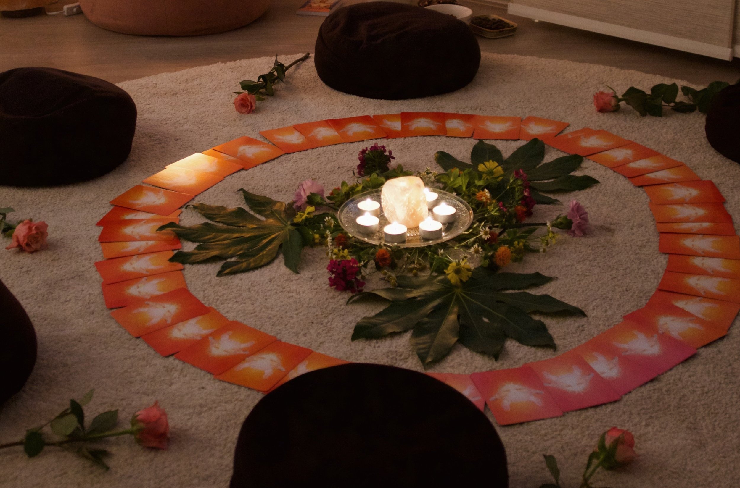 A circular arrangement of tarot cards on a rug with candles and leaves in the center, surrounded by cushions and flowers.
