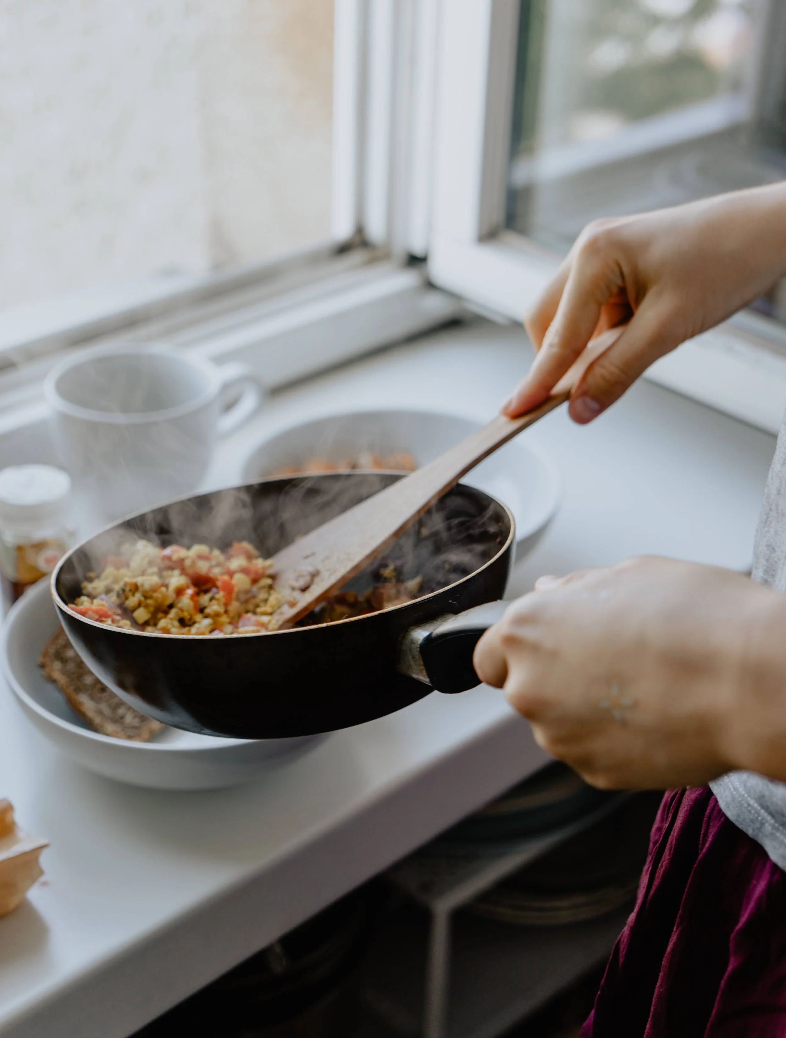 Person cooking food on a stovetop in a kitchen with a window in the background.