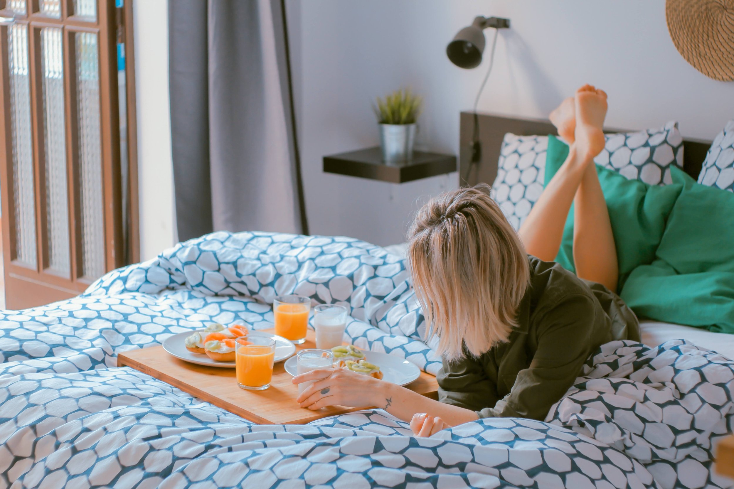 A woman with blonde hair lying on her stomach on a bed, reaching for a plate of breakfast food, with glasses of orange juice and a tray of food in front. Sunlight streams through a window, with a wall-mounted lamp, plant, and curtain decor visible in the background.