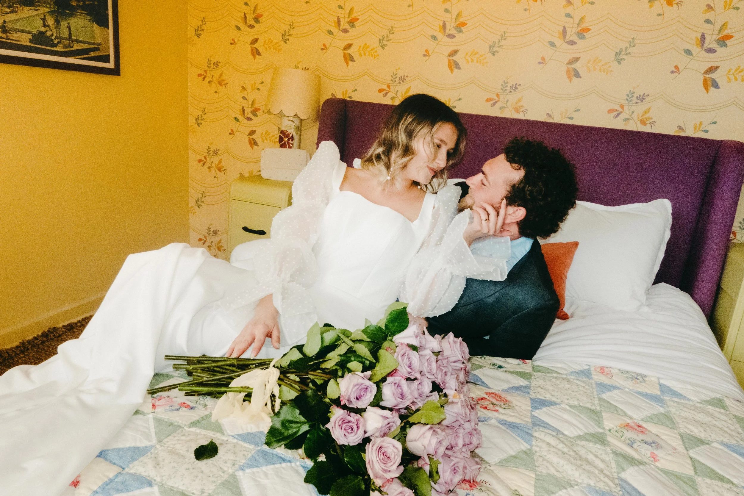 A woman in a white dress and a man in a suit lying on a bed, gazing at each other. There is a large bouquet of pink roses on the bed. The background shows a bedroom with a purple headboard, a nightstand with a lamp, and a wallpaper with floral patterns.