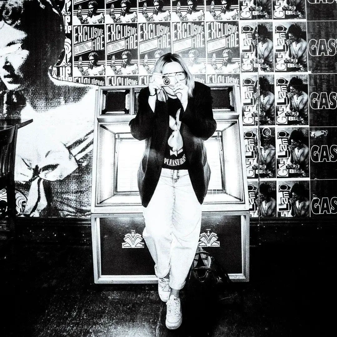 Photographer Alyssa Leicht taking a photo in front of a wall at the Gas Lamp music venue in Des Moines with various posters behind her. She is wearing a black jacket, a graphic T-shirt, and light-colored pants, standing in front of a jukebox.
