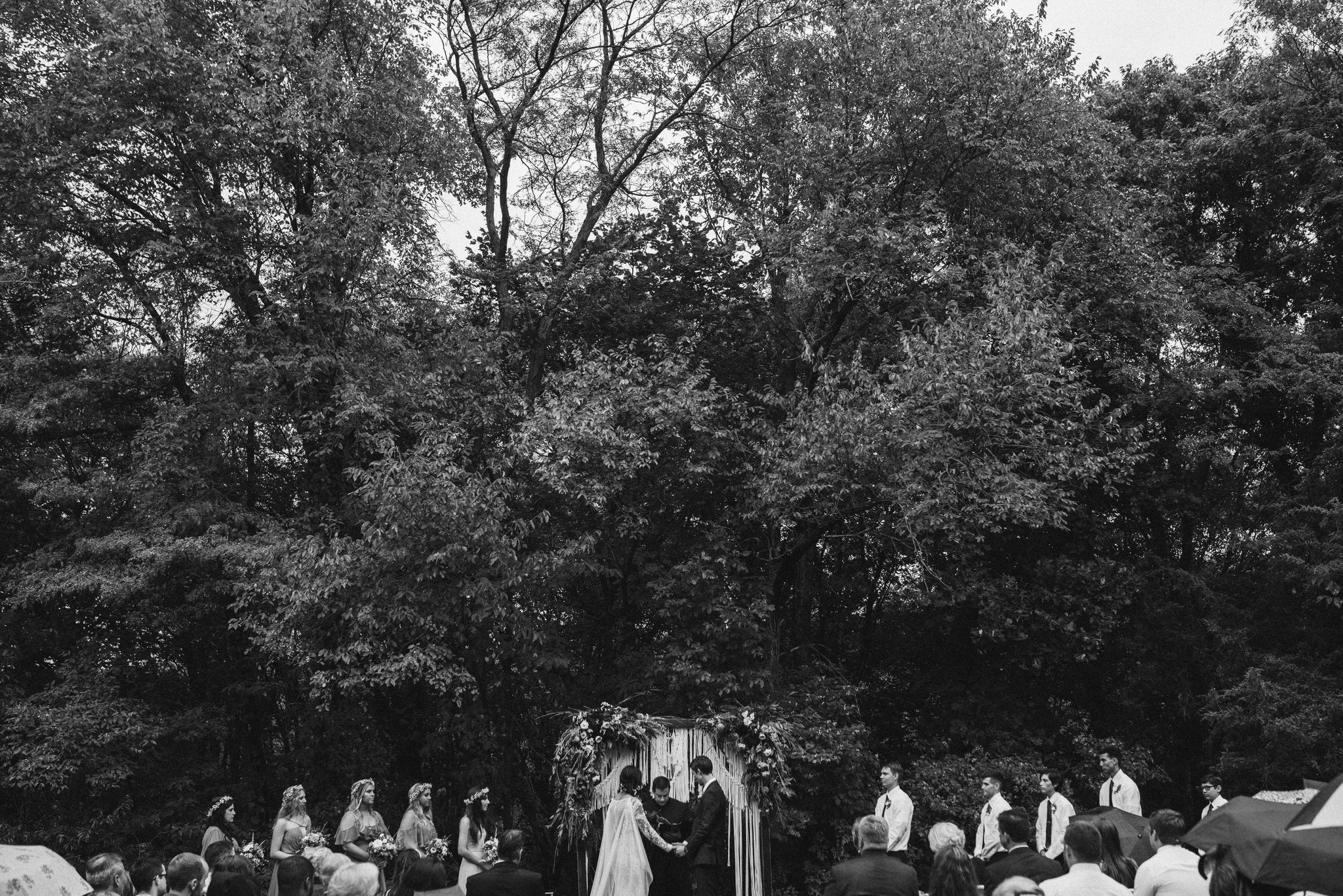 A black and white outdoor wedding ceremony at Jester Park in Iowa with a bride and groom standing under a decorated arch, surrounded by bridesmaids and groomsmen, with guests sitting and watching.