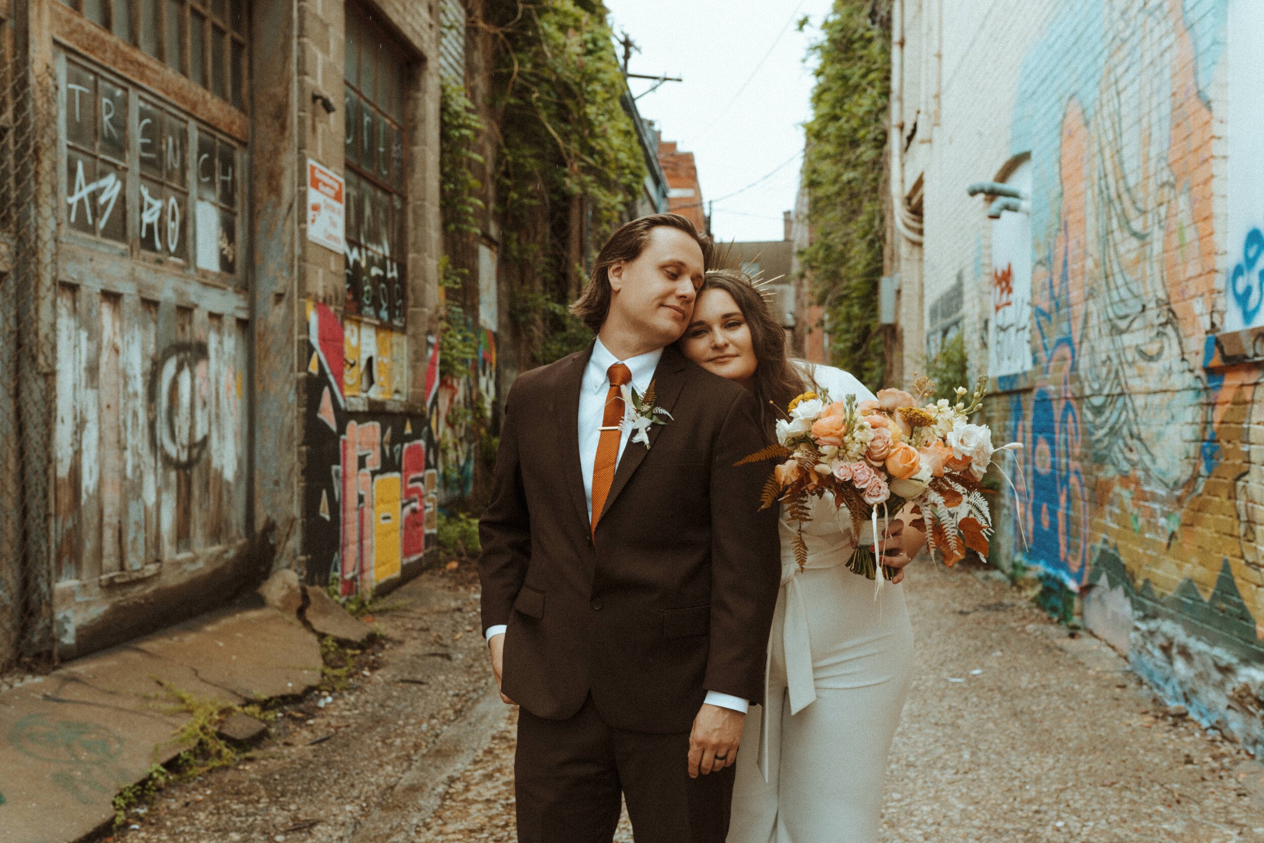 A couple in wedding attire walking hand in hand in an alleyway with graffiti-covered brick walls, the man in a dark suit and tie, and the woman holding a bouquet of flowers