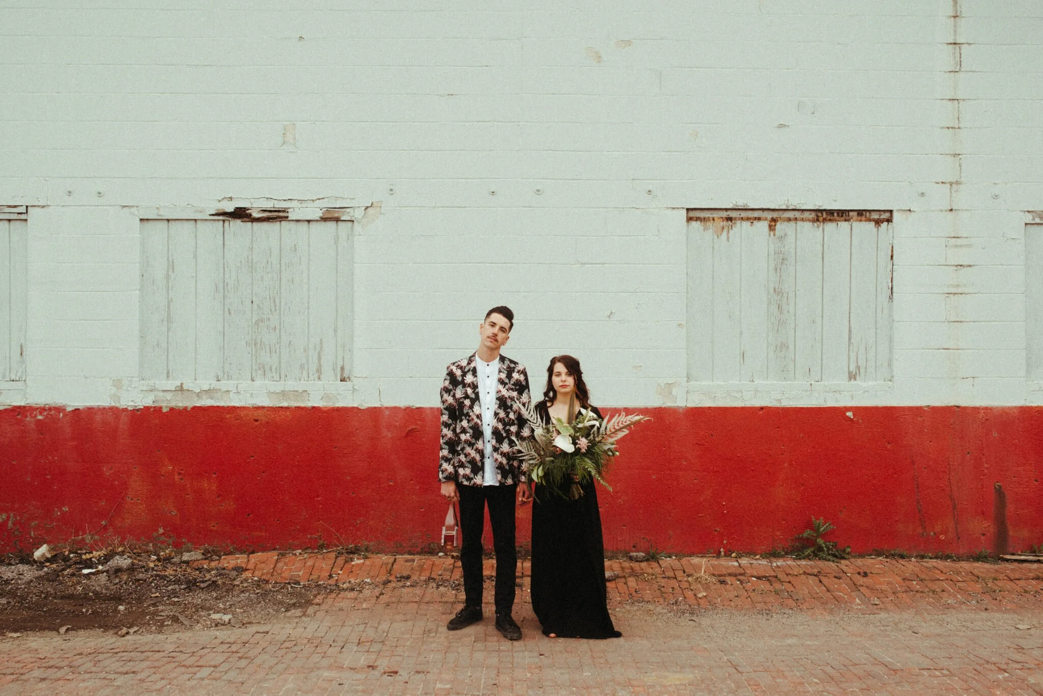 Two people, a man and a woman, standing against a large, weathered wall with peeling paint, holding a bouquet of flowers, with the man wearing a patterned blazer and the woman in a black dress with long hair.