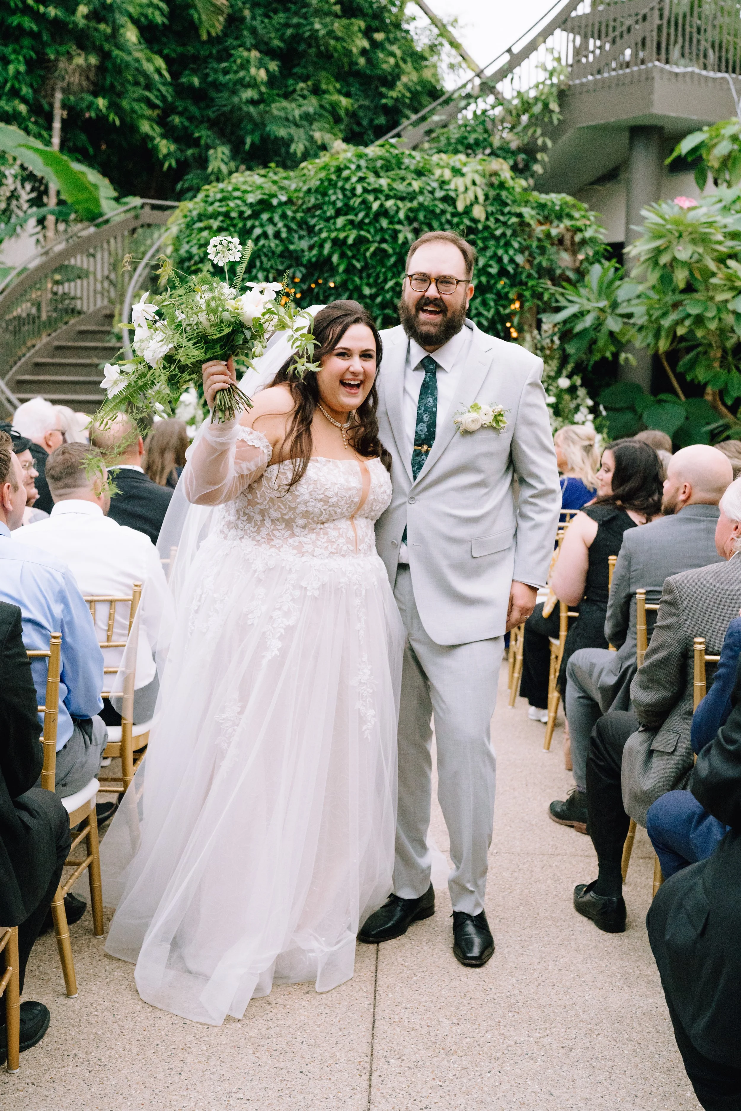 A bride and groom smiling and holding hands during their wedding ceremony at Greater Des Moines Botanical Gardens, with wedding guests seated around them outdoors.