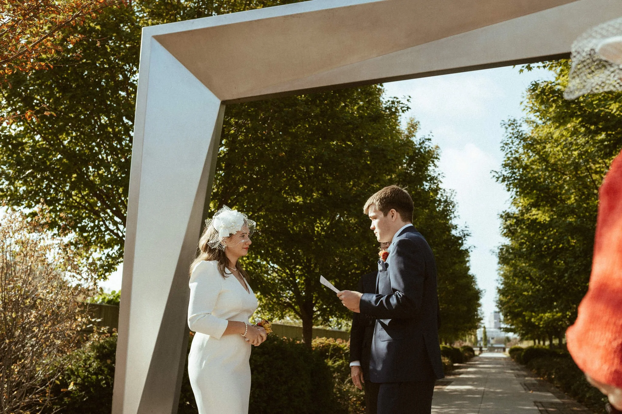 A couple getting married outdoors under a blue sky, with the bride wearing a white dress and veil, and the groom in a dark suit, standing in front of a white arch with leafy trees in the background.