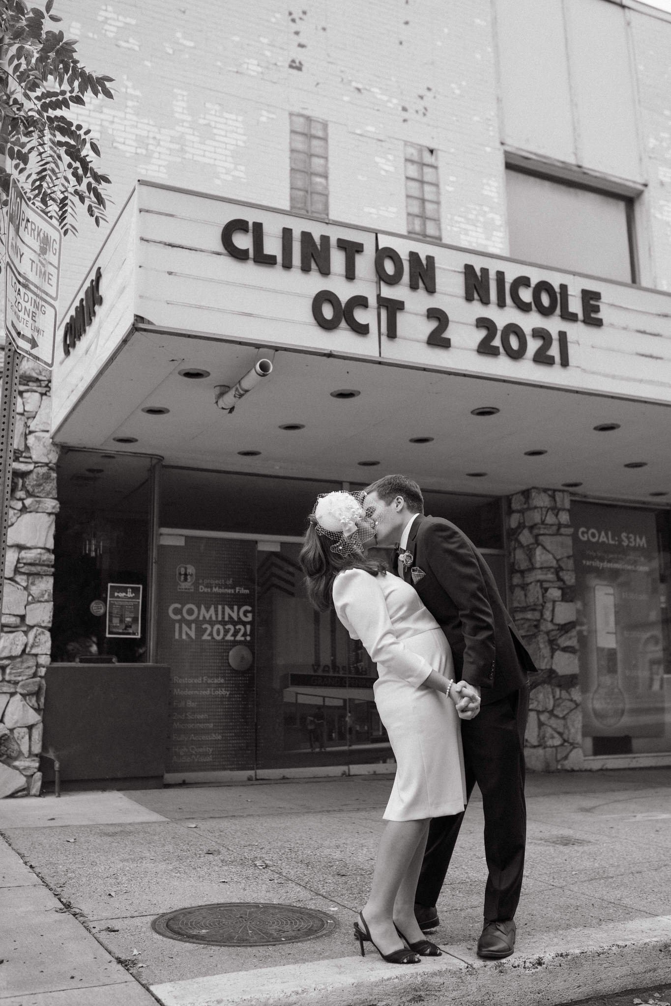 A couple dressed in formal attire sharing a kiss outside a theater with a marquee that reads "Clinton Nicole Oct 2 2021." The woman is wearing high heels and a white dress, and the man is in a dark suit. They are holding hands, standing close on a city sidewalk.
