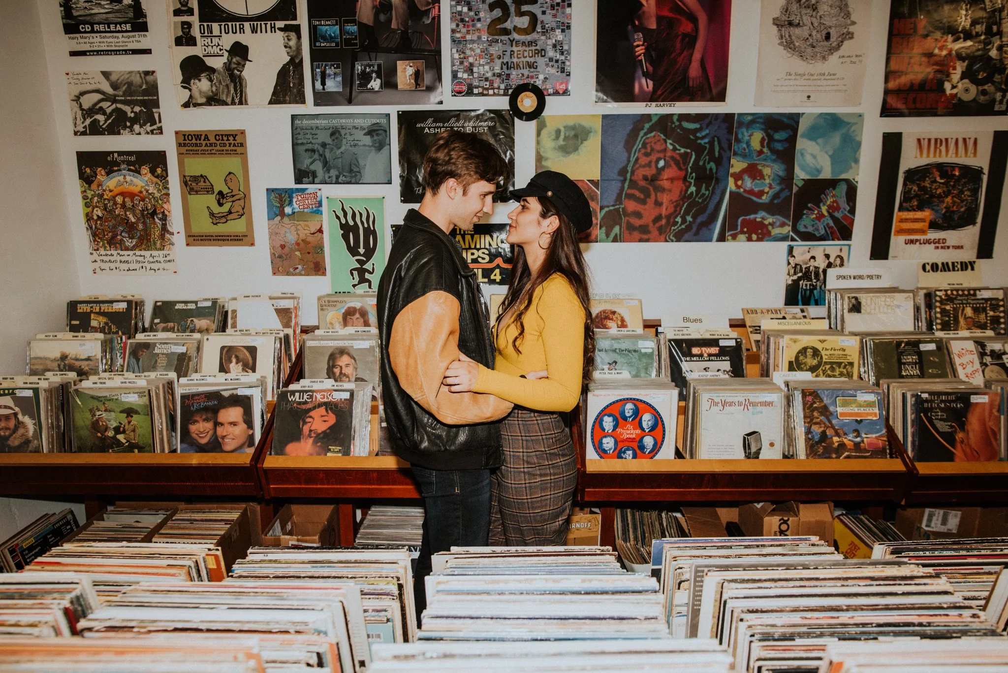 A young man and woman stand close together inside a record store, gazing into each other's eyes. The store's wall behind them is decorated with vintage posters, photographs, and album covers, creating a vibrant backdrop.