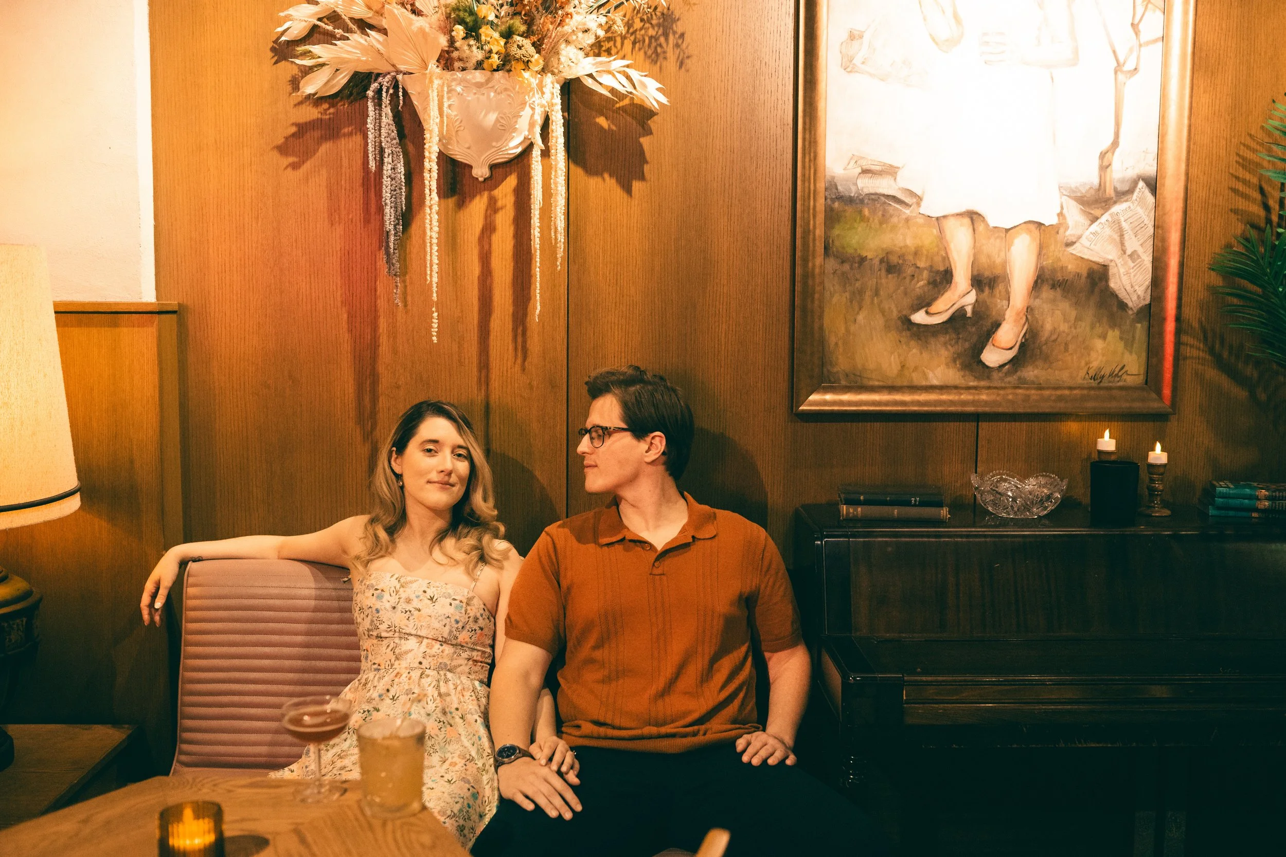 A young woman and a young man sitting together in a cozy, warmly lit room with wooden walls. The woman is wearing a floral dress and has long light brown hair, while the man is wearing a rust-colored polo shirt and glasses. There are three lit candles on the side table, and a painting of a woman in a white dress with high heels hanging on the wall behind them, with a large bouquet of dried flowers above the painting.