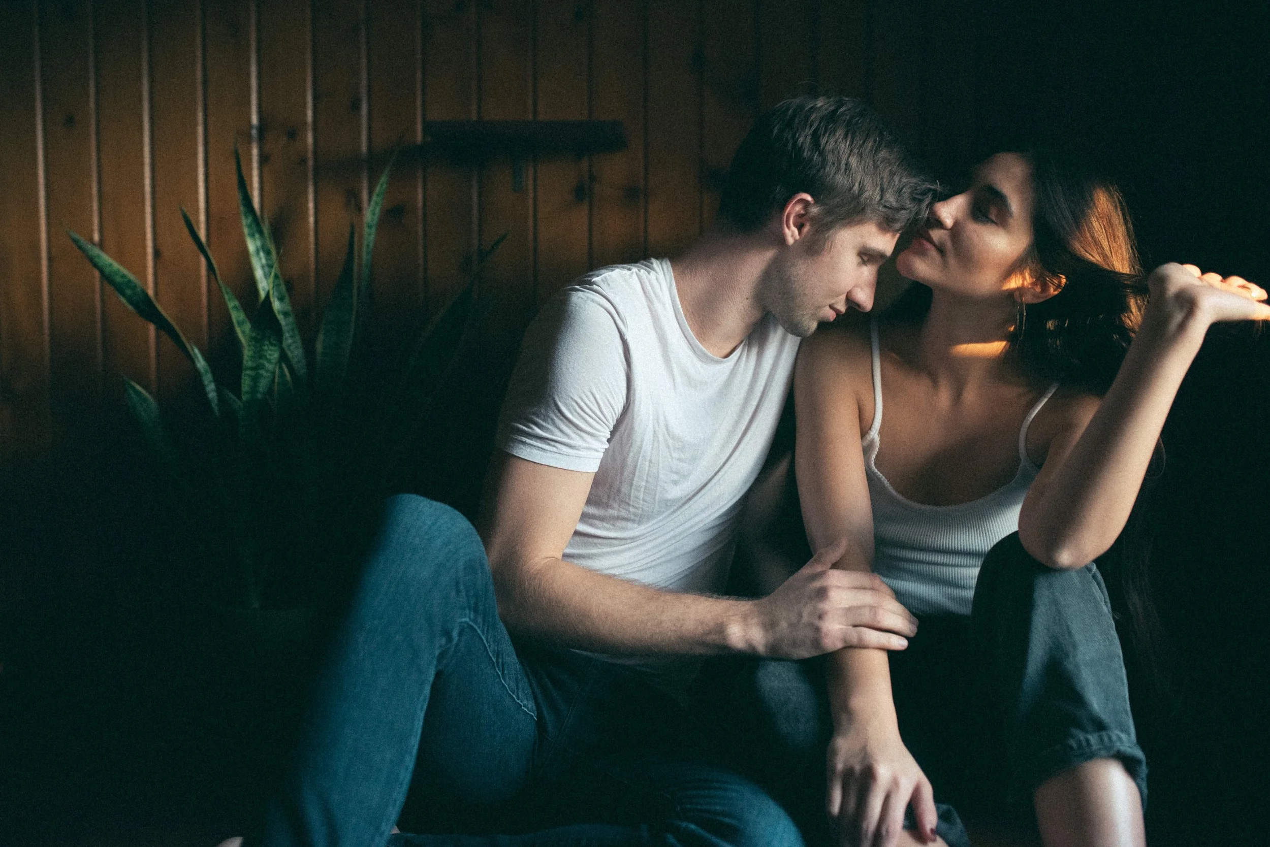 A young man and woman sitting closely together, touching foreheads, in a dimly lit room with wood-paneled walls and a plant in the background.