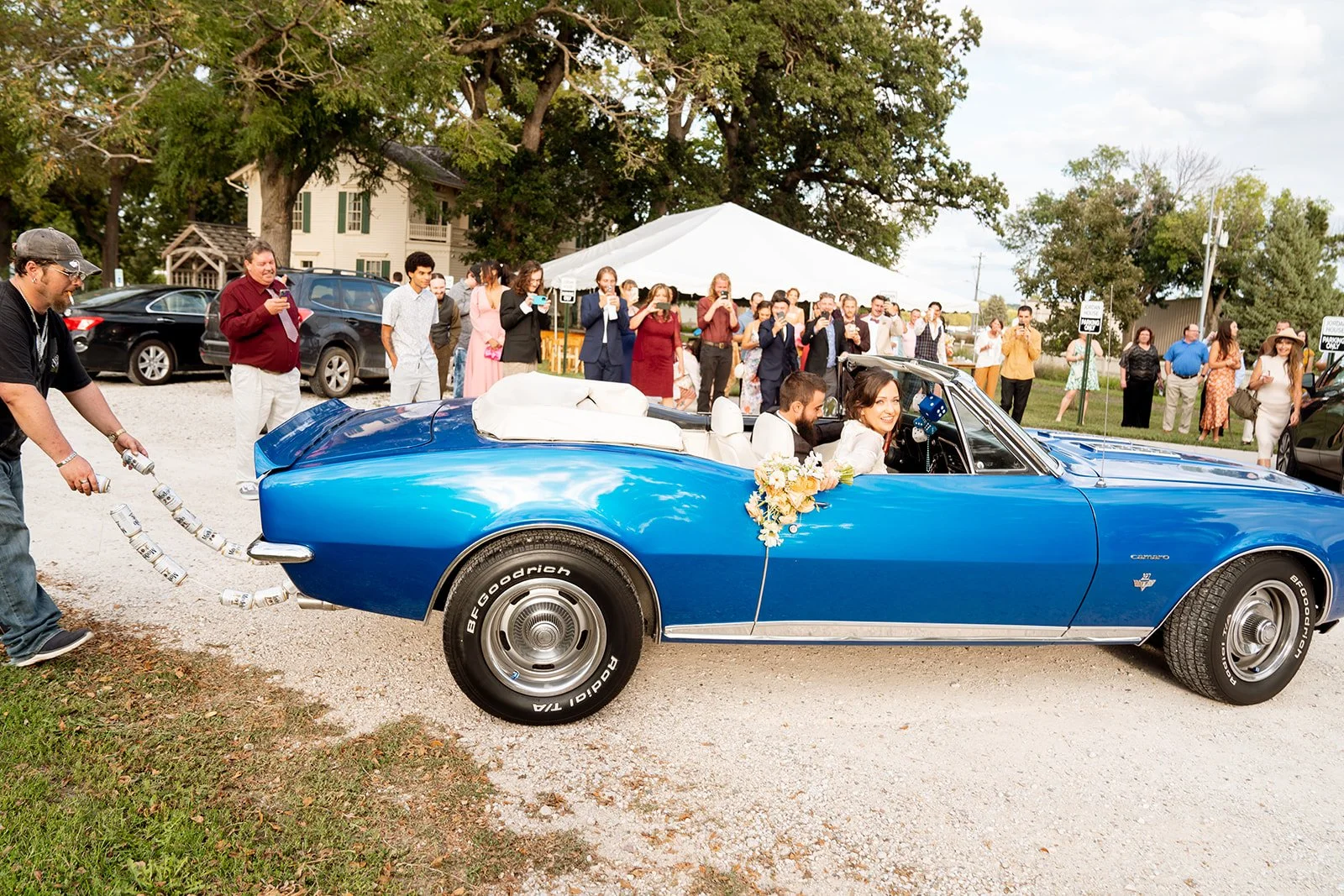 A bride and groom sitting in a vintage blue convertible car, with the bride holding a bouquet of flowers, at a wedding celebration. Guests are taking photos in the background, some under a white tent and some standing outdoors.