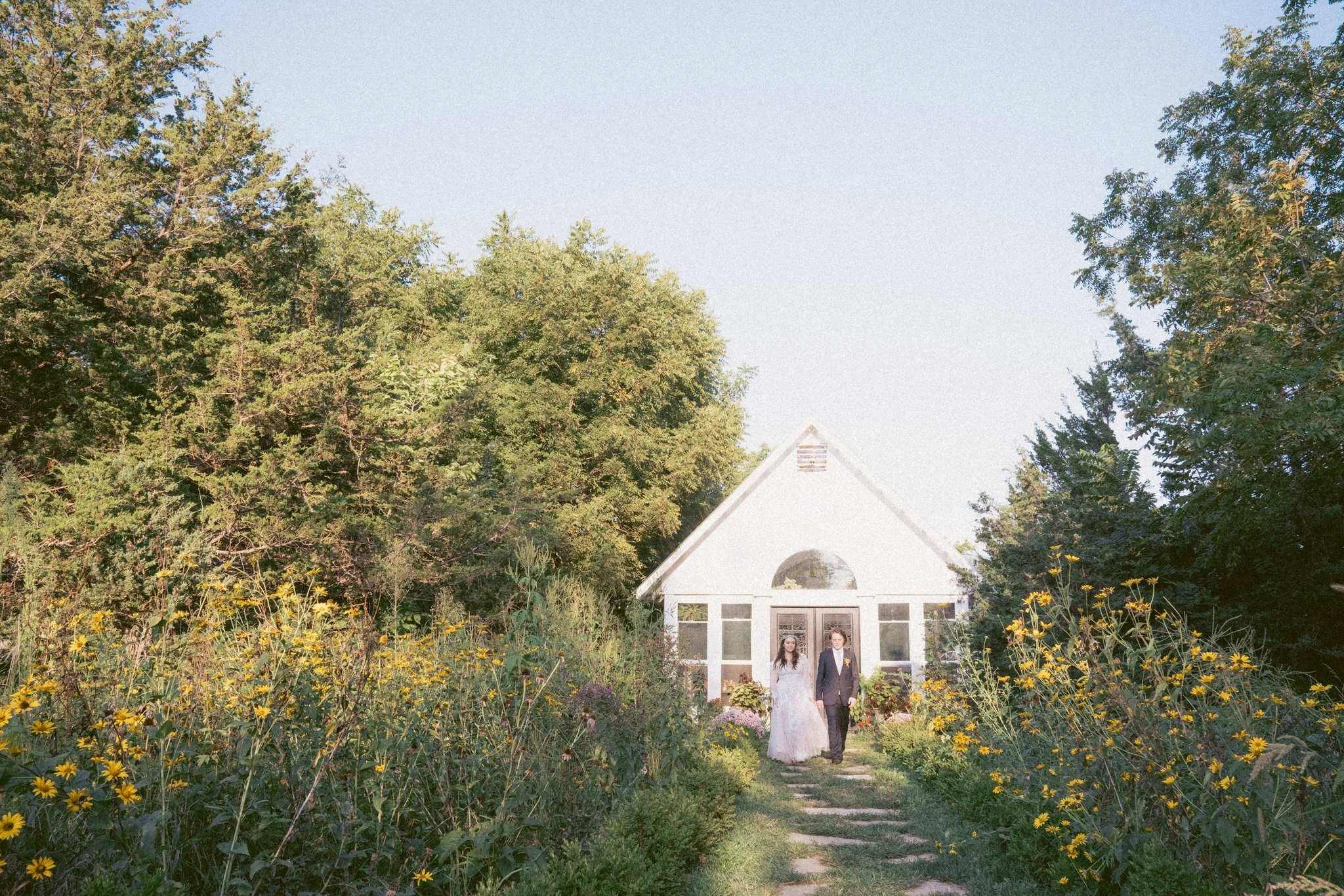 A bride and groom walking down a garden path towards a white greenhouse with large windows and a triangular roof, surrounded by tall green trees and yellow flowers. At Iris Aisle wedding venue in Winterset.