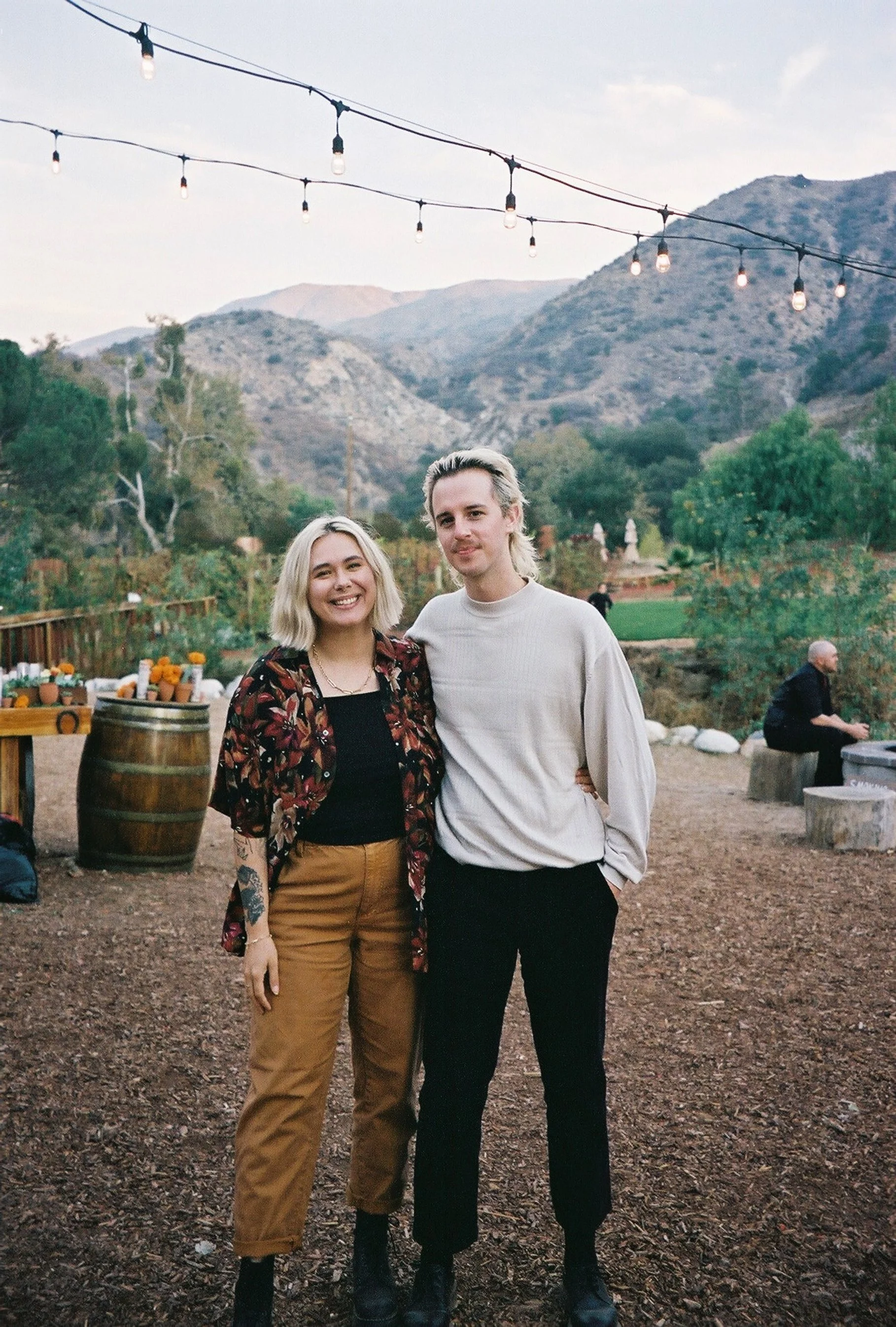 Traveling wedding photographer Alyssa Leicht and her partner, standing outside at a wedding venue in a scenic mountainous area in Los Angeles with string lights overhead.
