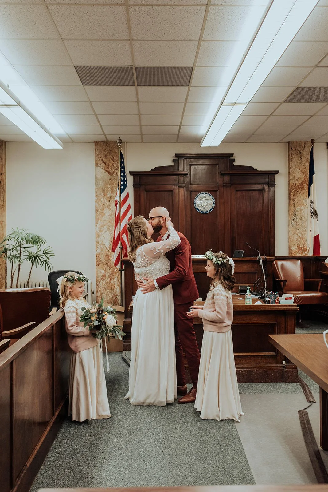 A bride and groom kiss in a courtroom, with two young girls dressed in white skirts and blush pink tops, one holding a bouquet of flowers, all standing in front of wooden judge's bench, with American and California flags in the background.