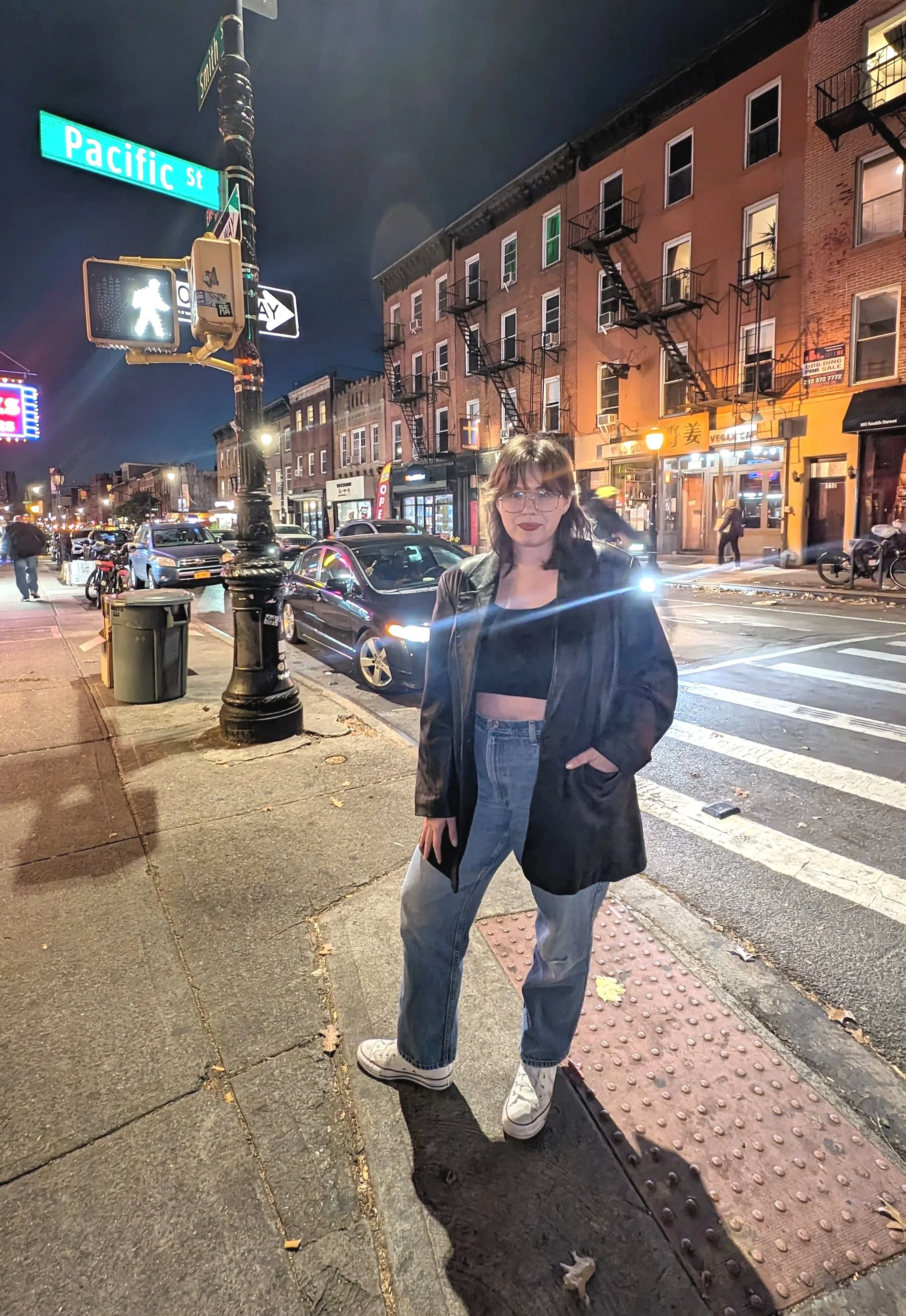 Nighttime street scene showing a woman standing on a city sidewalk at an intersection near Pacific Street. She has dark hair and glasses, is wearing a black top and jacket, light blue jeans, and white sneakers. The background features cars, brick buildings, storefronts, and a person riding a bike.