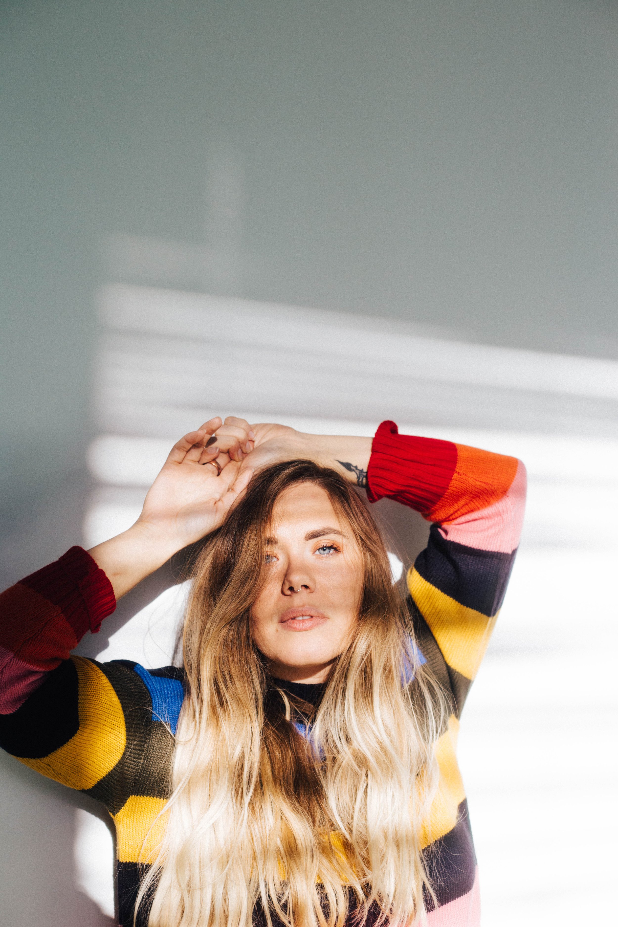 A woman with long wavy blonde hair and blue eyes, wearing a colorful striped sweater, poses with her hands on her head against a background of natural light and shadows.