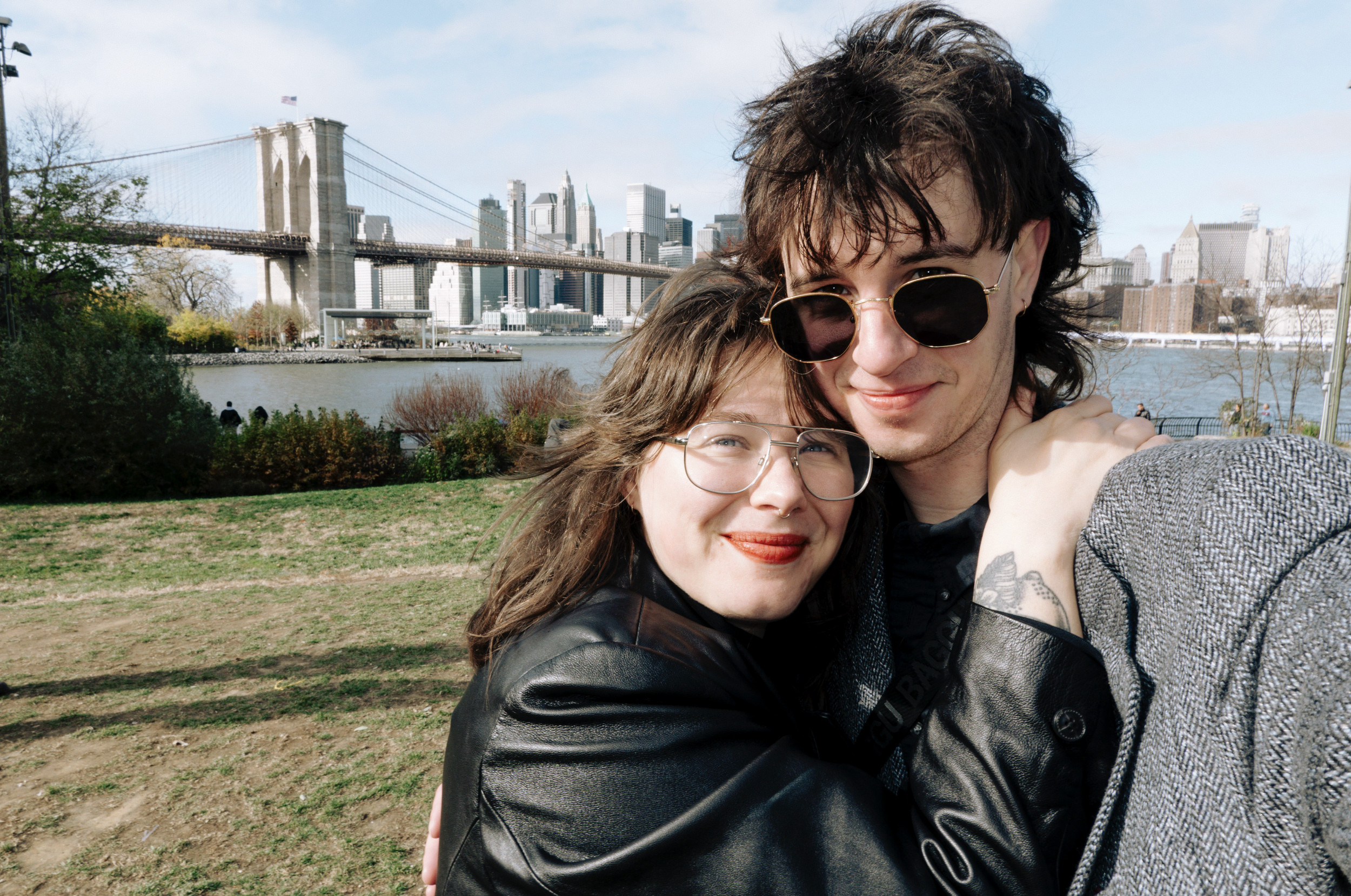 A young couple taking a selfie in a park with the New York City skyline and Brooklyn Bridge in the background.