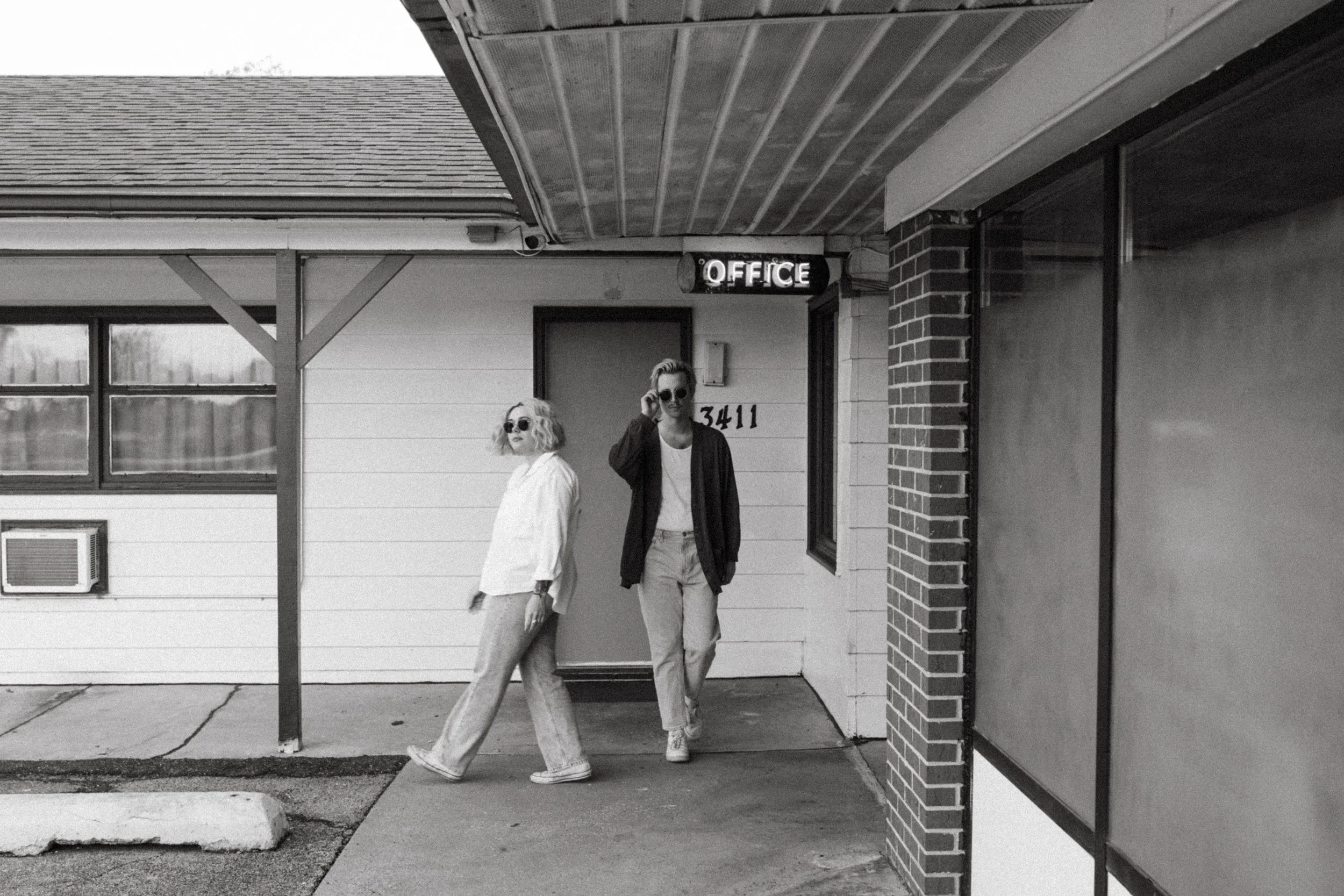 A black and white photo of a couple standing outside in front of an abandoned roadside motel. The man is adjusting his sunglasses while the woman walks across the scene.