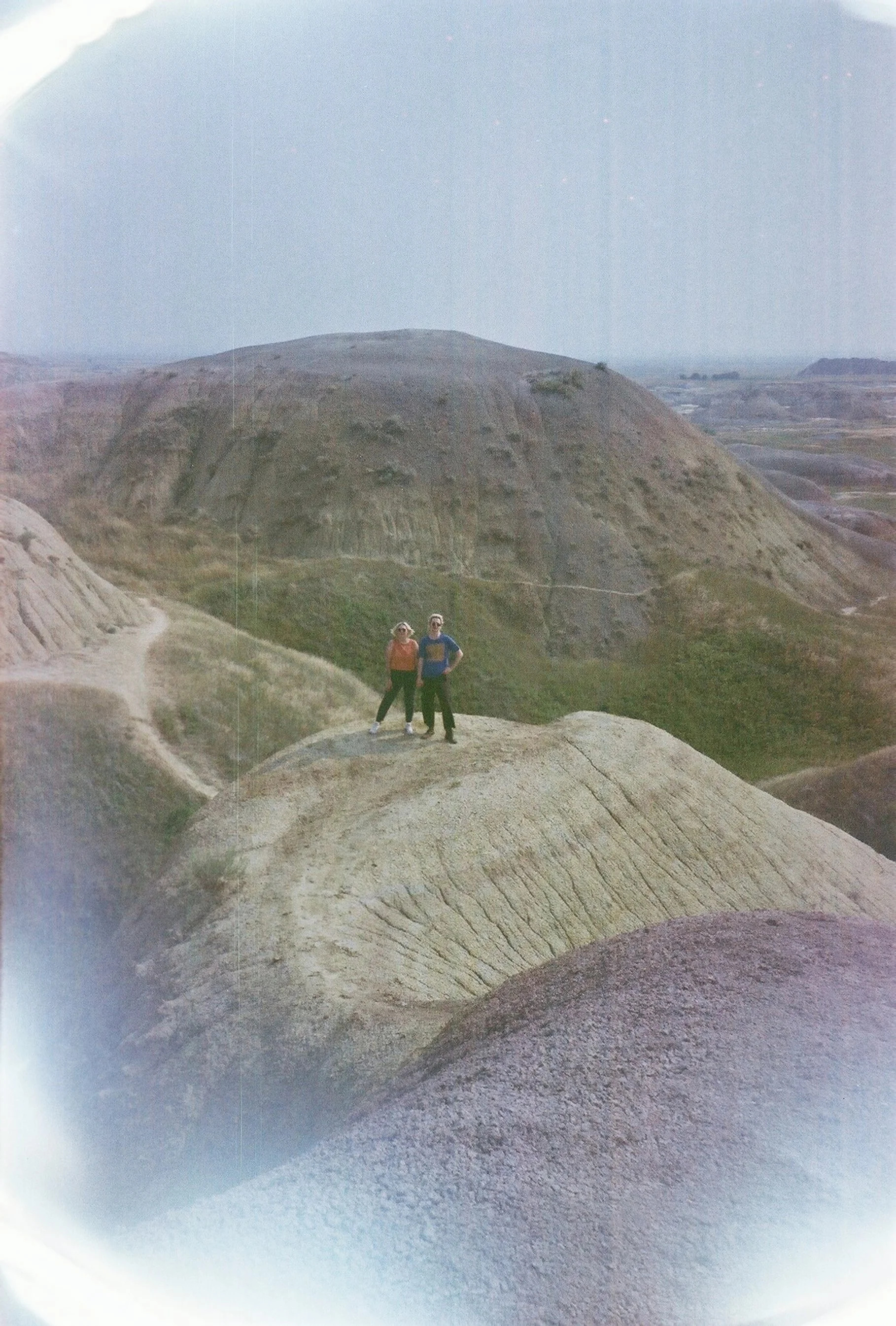 Two people standing on a large, light-colored rock formation in a hilly landscape with sparse vegetation and a valley in the background.