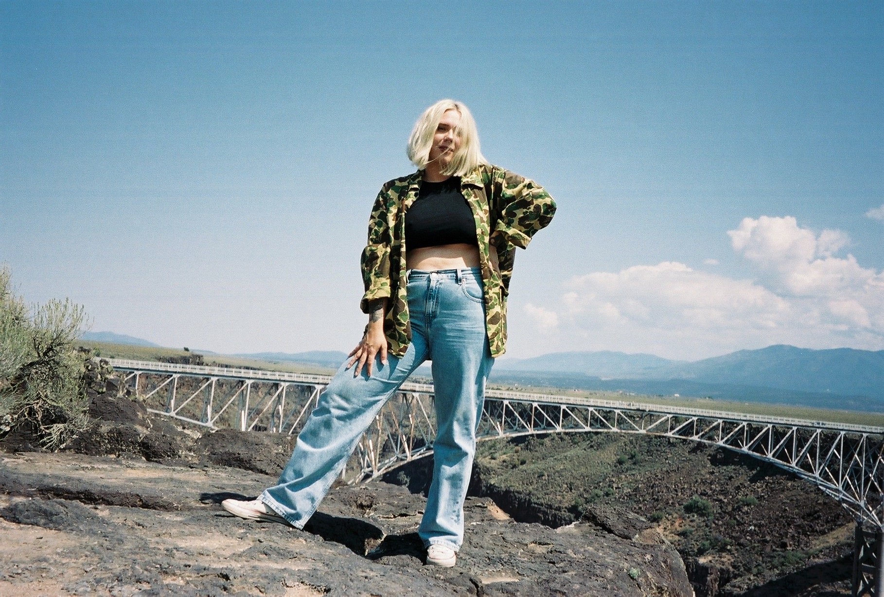 A woman with blonde hair posing outdoors on rocky terrain with a bridge and mountains in the background, wearing a camouflage jacket, black crop top, and light blue jeans.