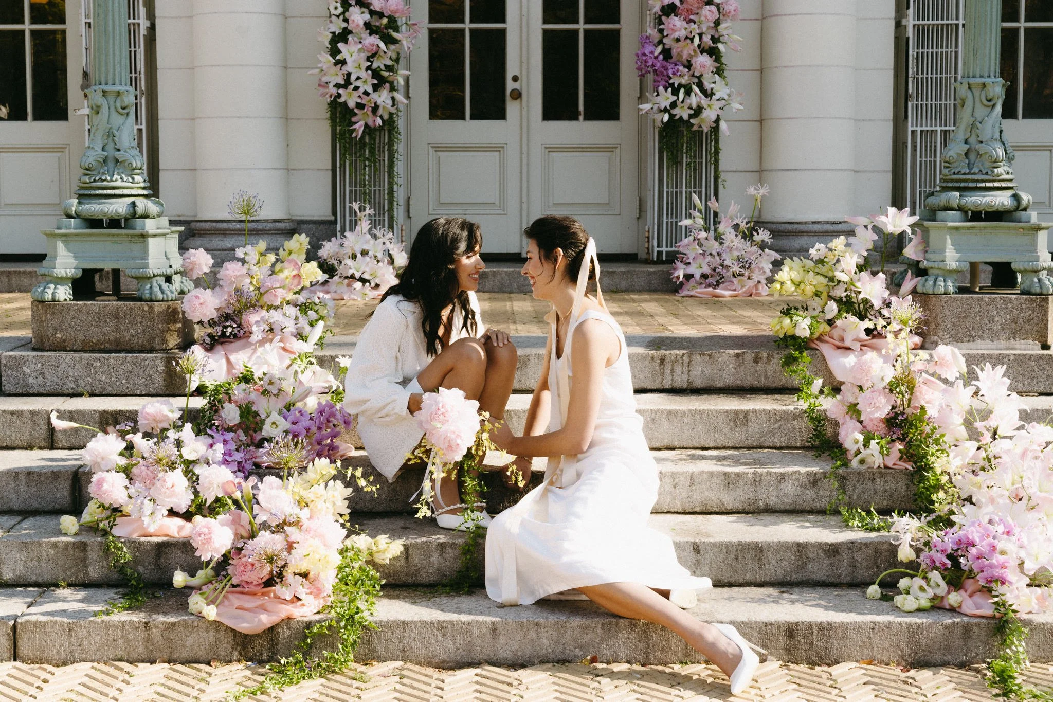 Lesbian brides sitting on steps in front of a white door decorated with pink and white flowers, surrounded by bouquets of similar flowers. wlw elopement in Brooklyn New York.