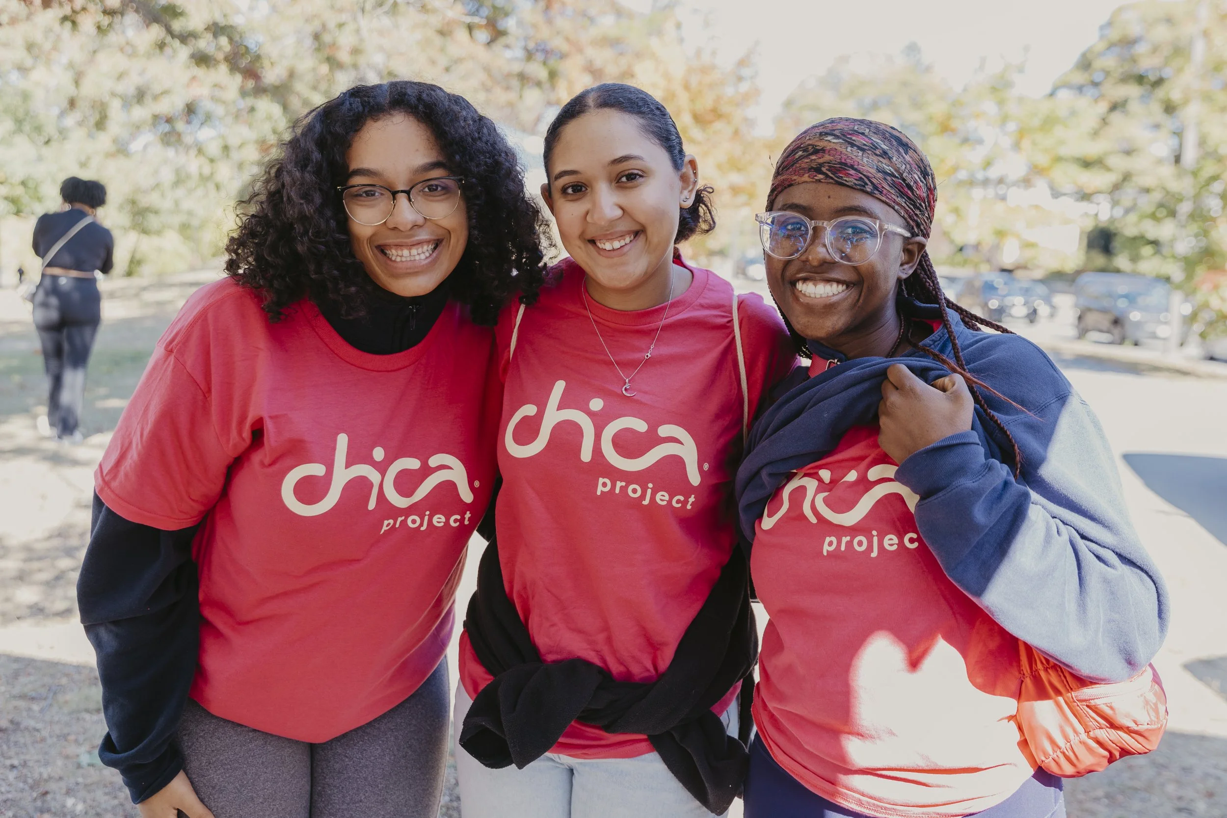 Three Chica Project participants smiling together outdoors in Boston wearing Chica Project t-shirts, celebrating sisterhood and community among young Women of Color