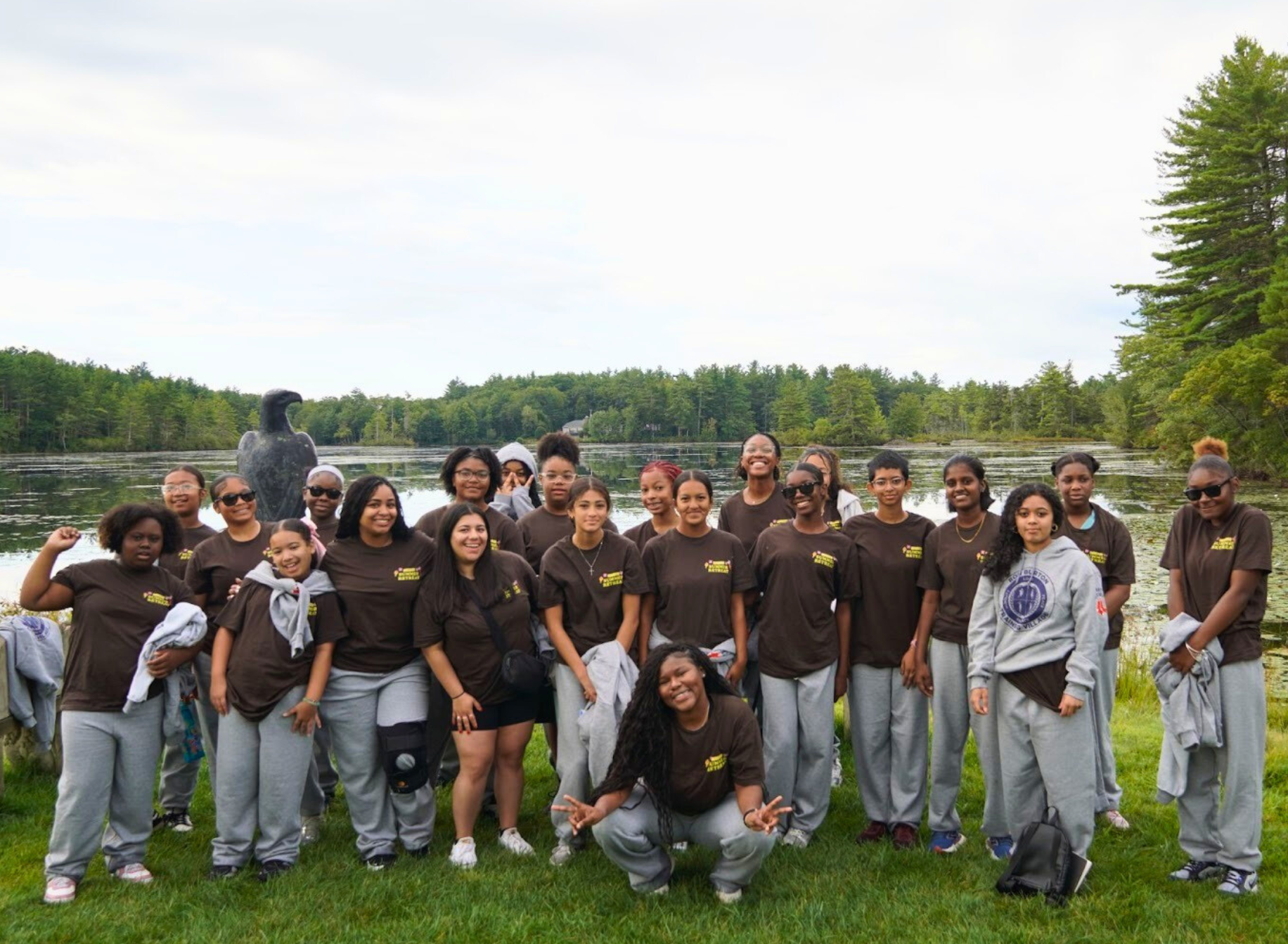 A group of young women and girls standing by a pond outdoors, some wearing matching brown shirts, posing for a photo with trees and a shoreline in the background.