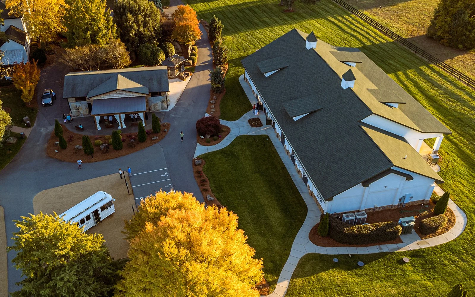 Aerial view of a large building and adjacent smaller structure in a landscaped area with green lawns and trees with autumn foliage. A vehicle is parked nearby, and several people are visible outside the structures.