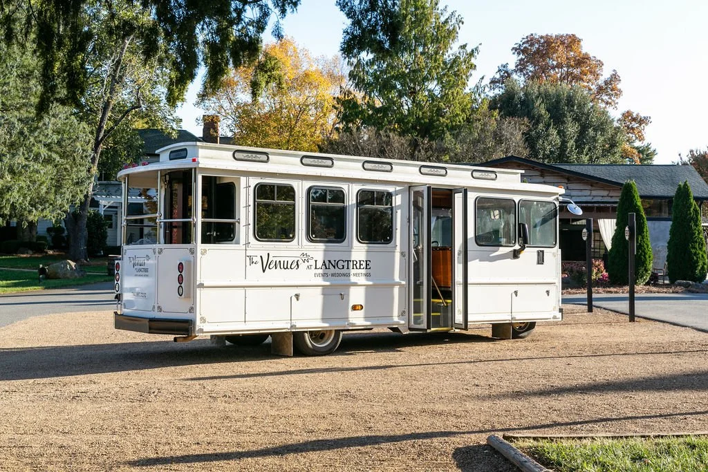 White event trolley bus parked on a gravel pathway near trees and a building.