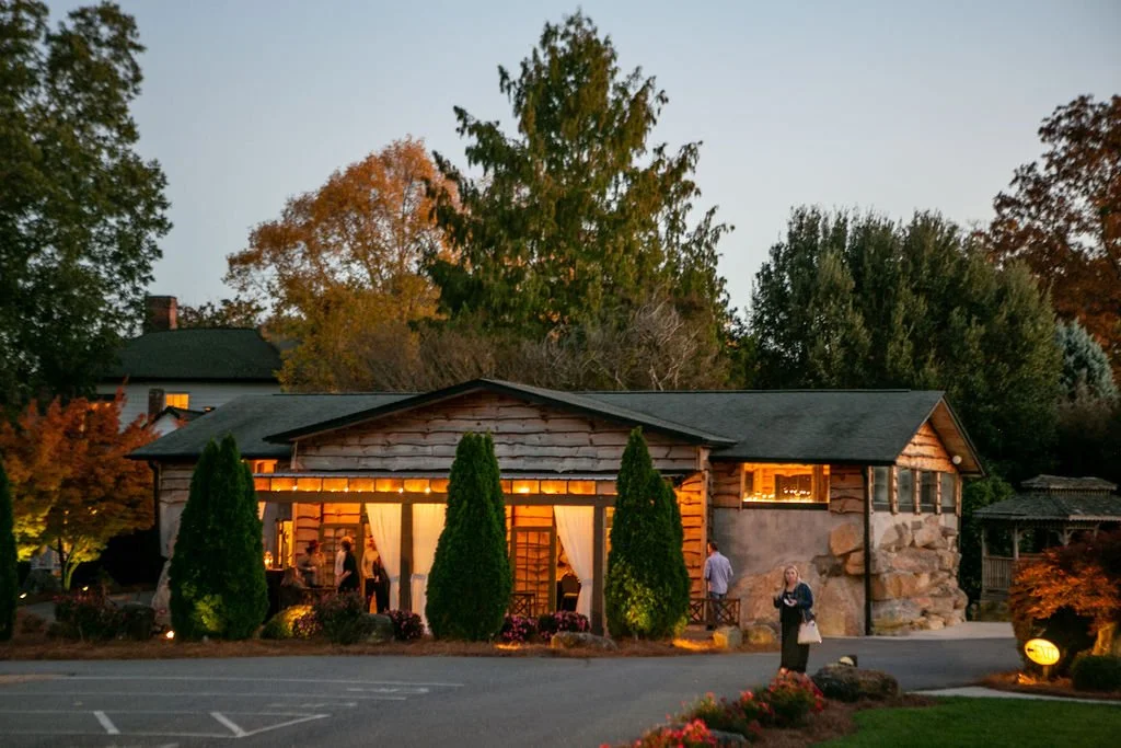 Rustic wooden building with lit interior at dusk, surrounded by trees and shrubs, people nearby, under a clear evening sky.