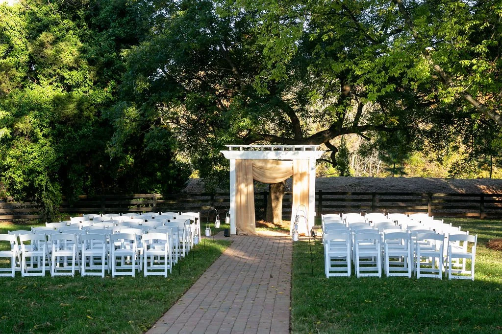Outdoor wedding ceremony setup with a wooden pergola draped in fabric, surrounded by white chairs on a grass lawn, with trees and a wooden fence in the background.