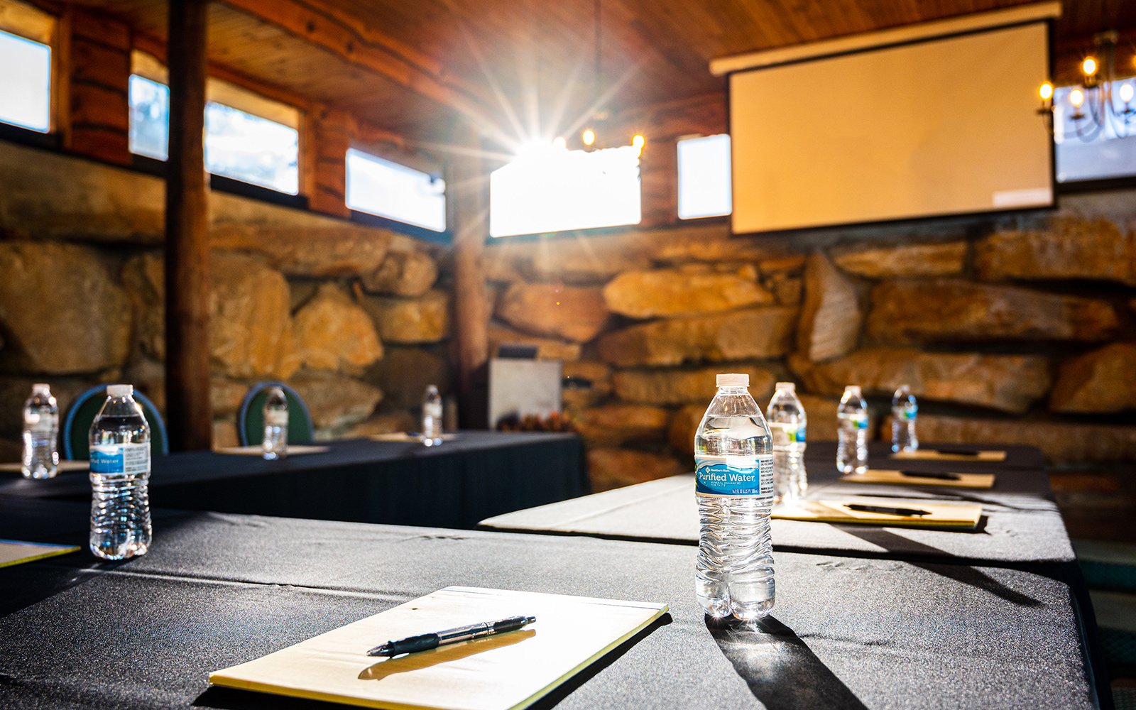 Conference room with water bottles and notepads on tables, projector screen in background, natural stone wall, and sunlight streaming in.