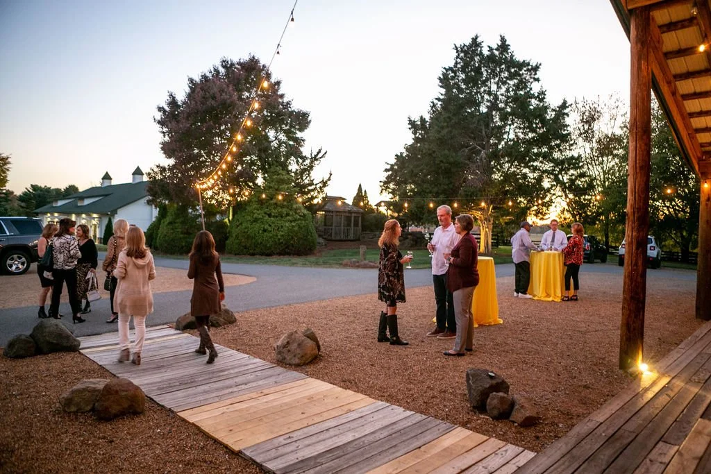 People gathered outdoors at an event, standing near tables with yellow tablecloths, surrounded by trees and string lights at dusk.