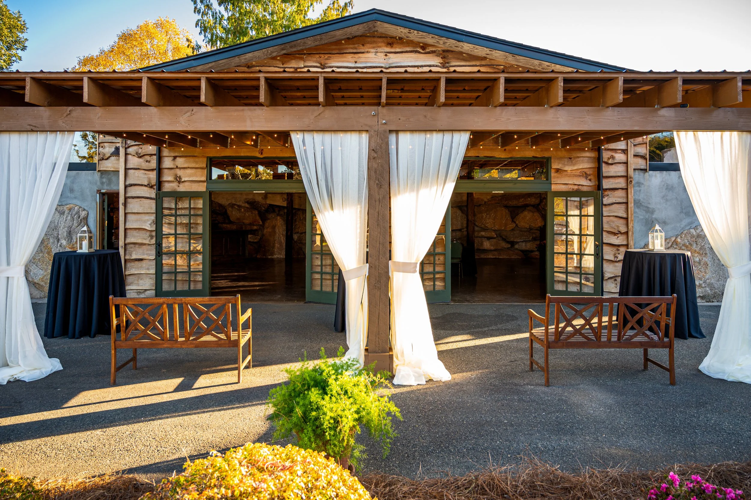 An outdoor wooden structure with white drapes, wooden benches, and tables with lanterns. The structure has open doors and a stone wall visible inside. The area is decorated with plants and lit by natural sunlight.