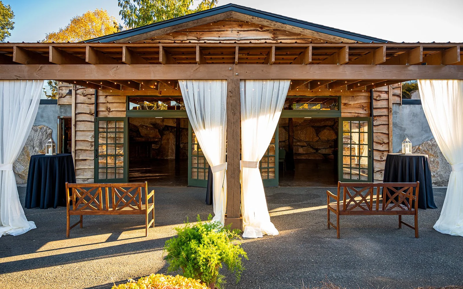 Rustic wooden pavilion with white curtains, wooden benches, and small round tables with lanterns, set outdoors with autumn foliage background.