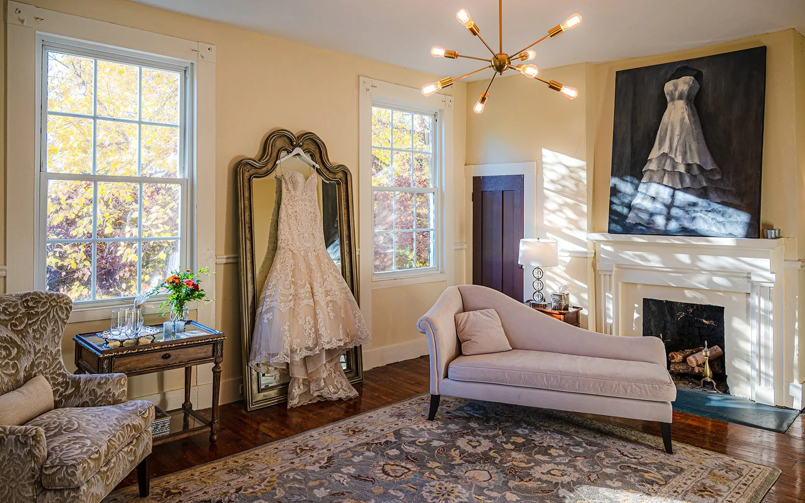 Elegant room with a wedding gown hanging on a vintage mirror, chaise lounge, armchair, fireplace, and artwork of a dress.