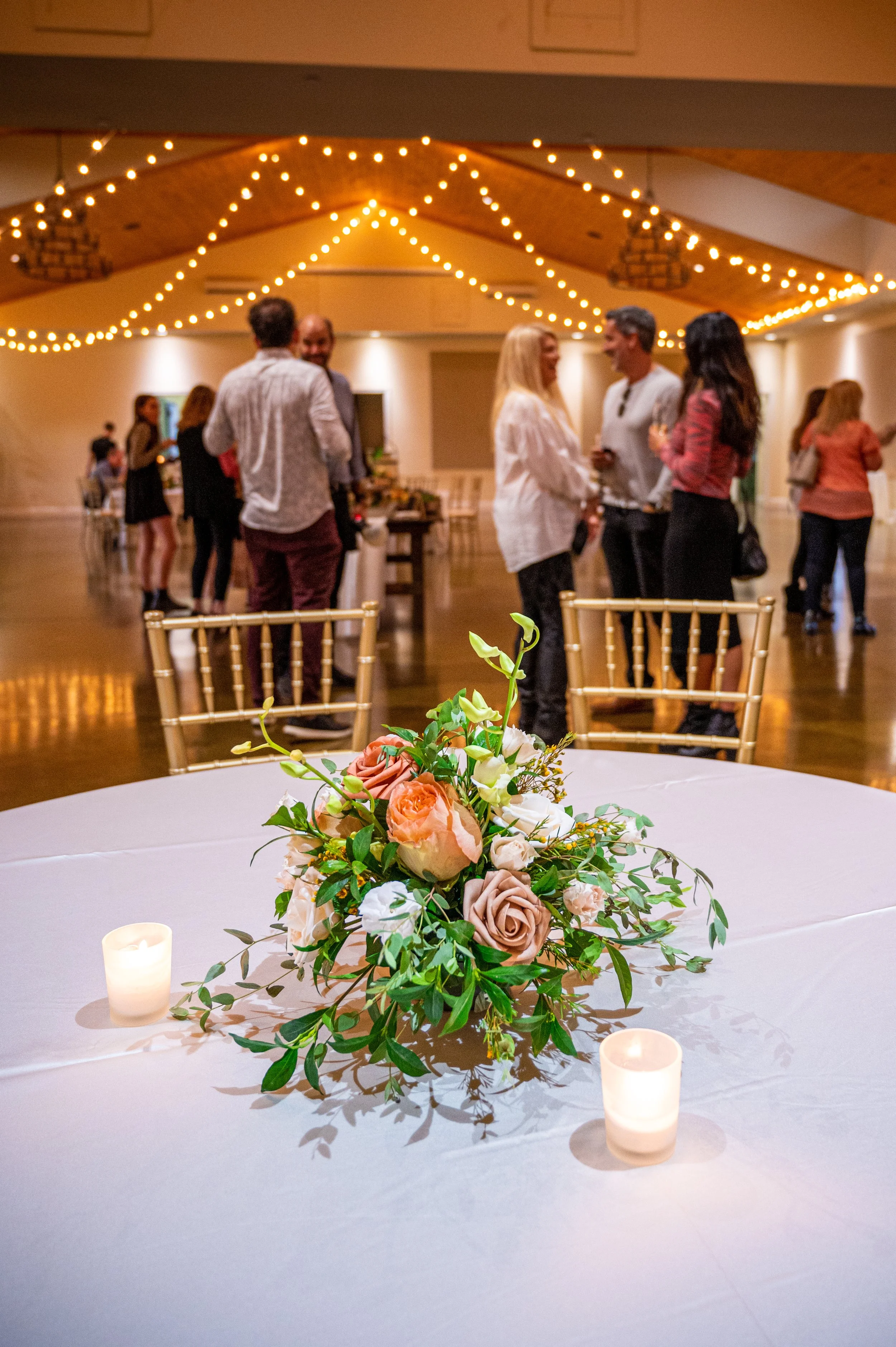 Event hall with a decorated table featuring a floral centerpiece and candles in the foreground, with people mingling under string lights in the background.