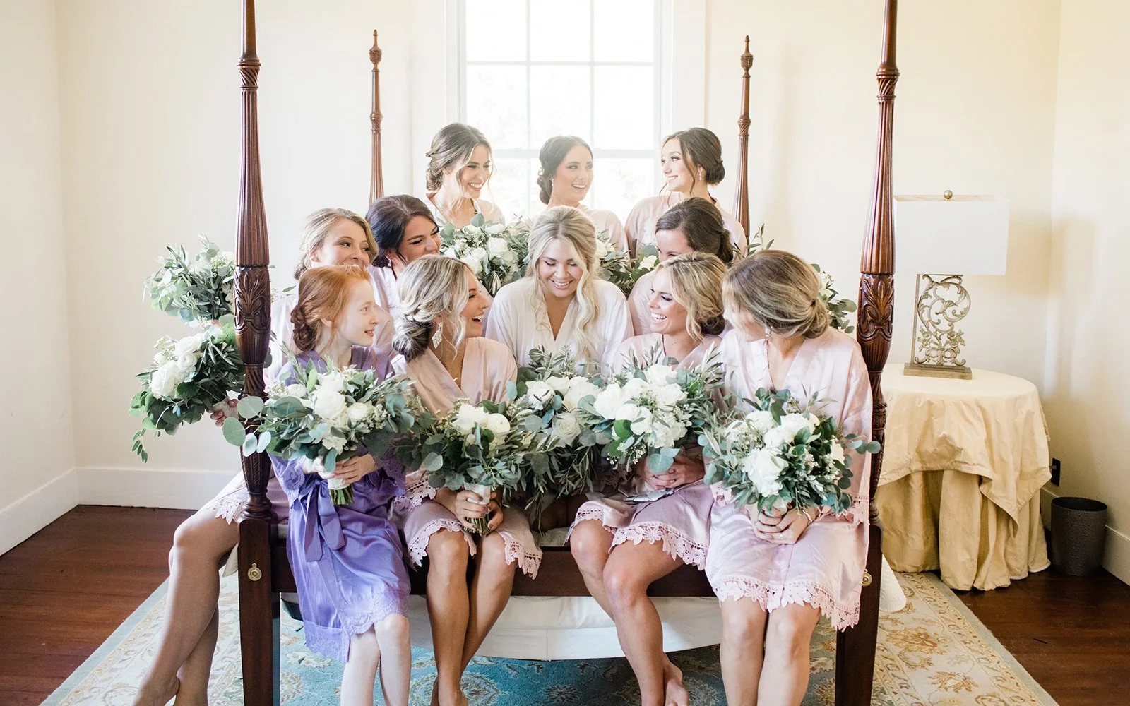 Bridal party sitting on bed, holding bouquets, wearing robes, smiling.