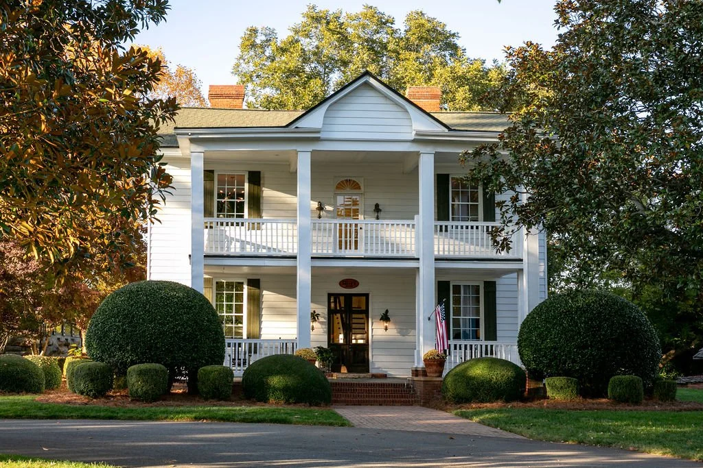 Two-story white colonial house with a porch and columns, surrounded by manicured shrubs and trees.