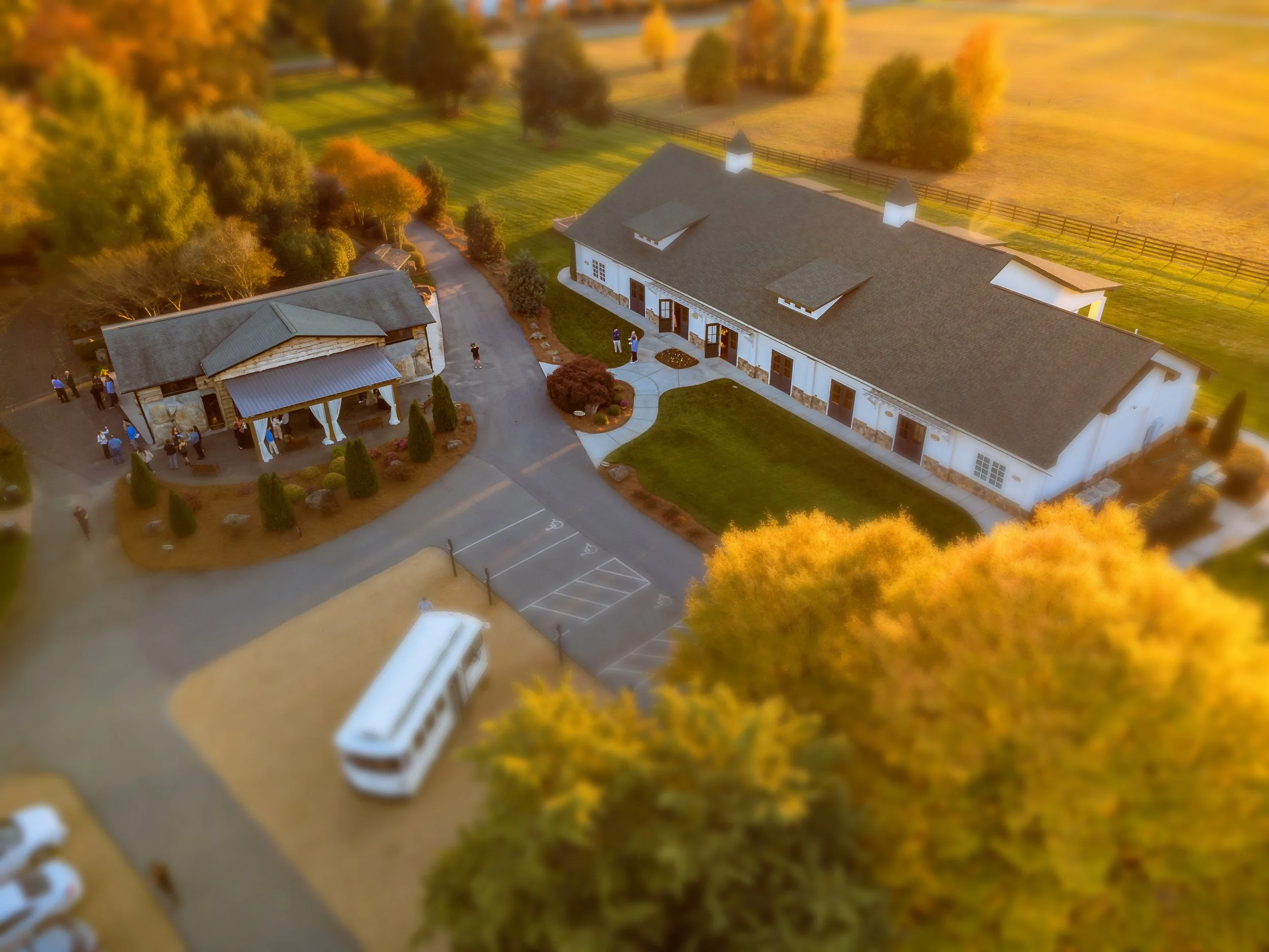 Aerial view of a wedding venue with a large white building and another smaller building, surrounded by trees and open fields, a bus is parked nearby, and people are gathered outside, captured in the fall season with orange and golden foliage.