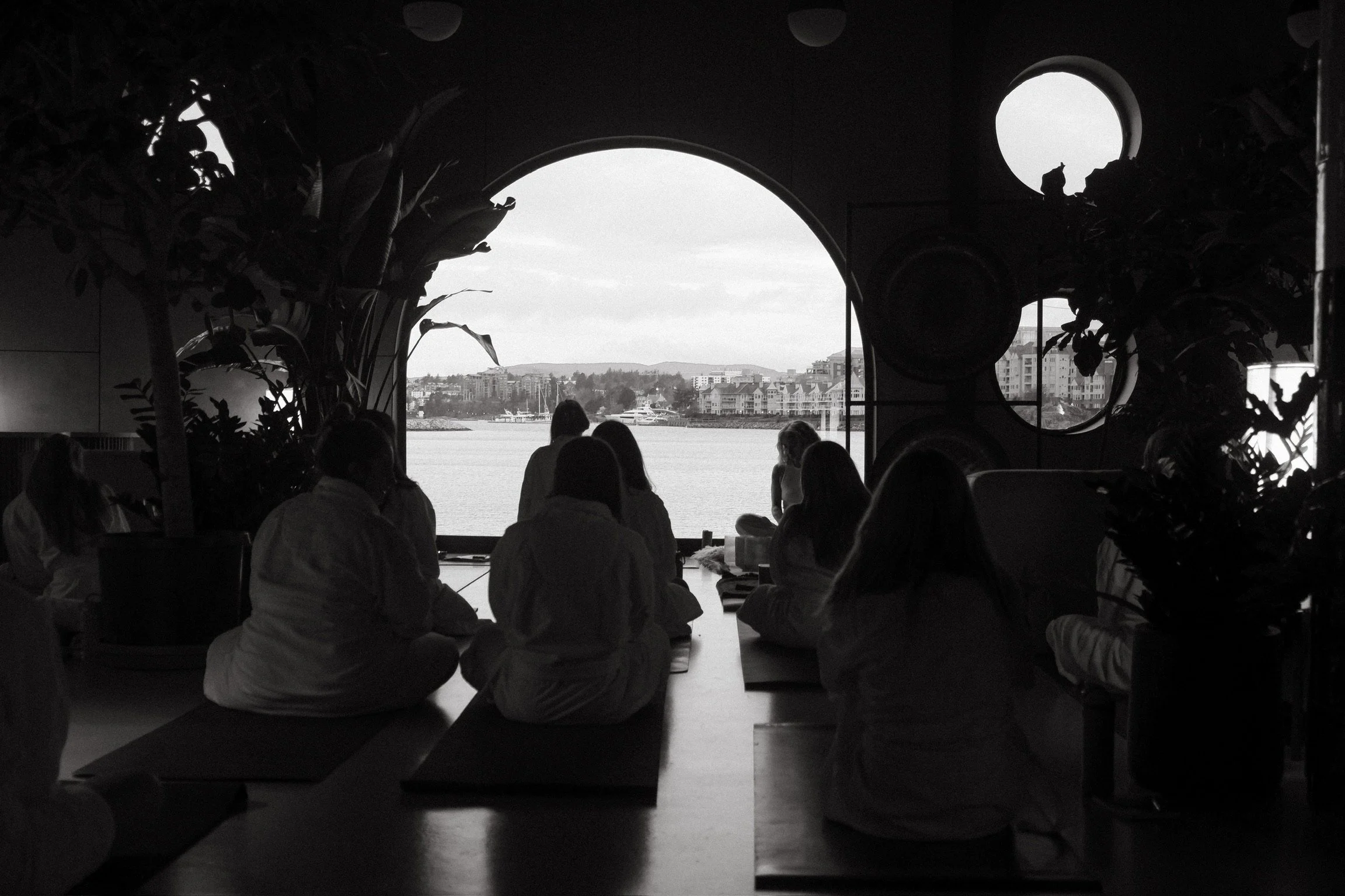 Silhouetted group of people sitting on mats in a meditation or yoga class, facing a large window overlooking a body of water and cityscape, with interior plants and round decorations.