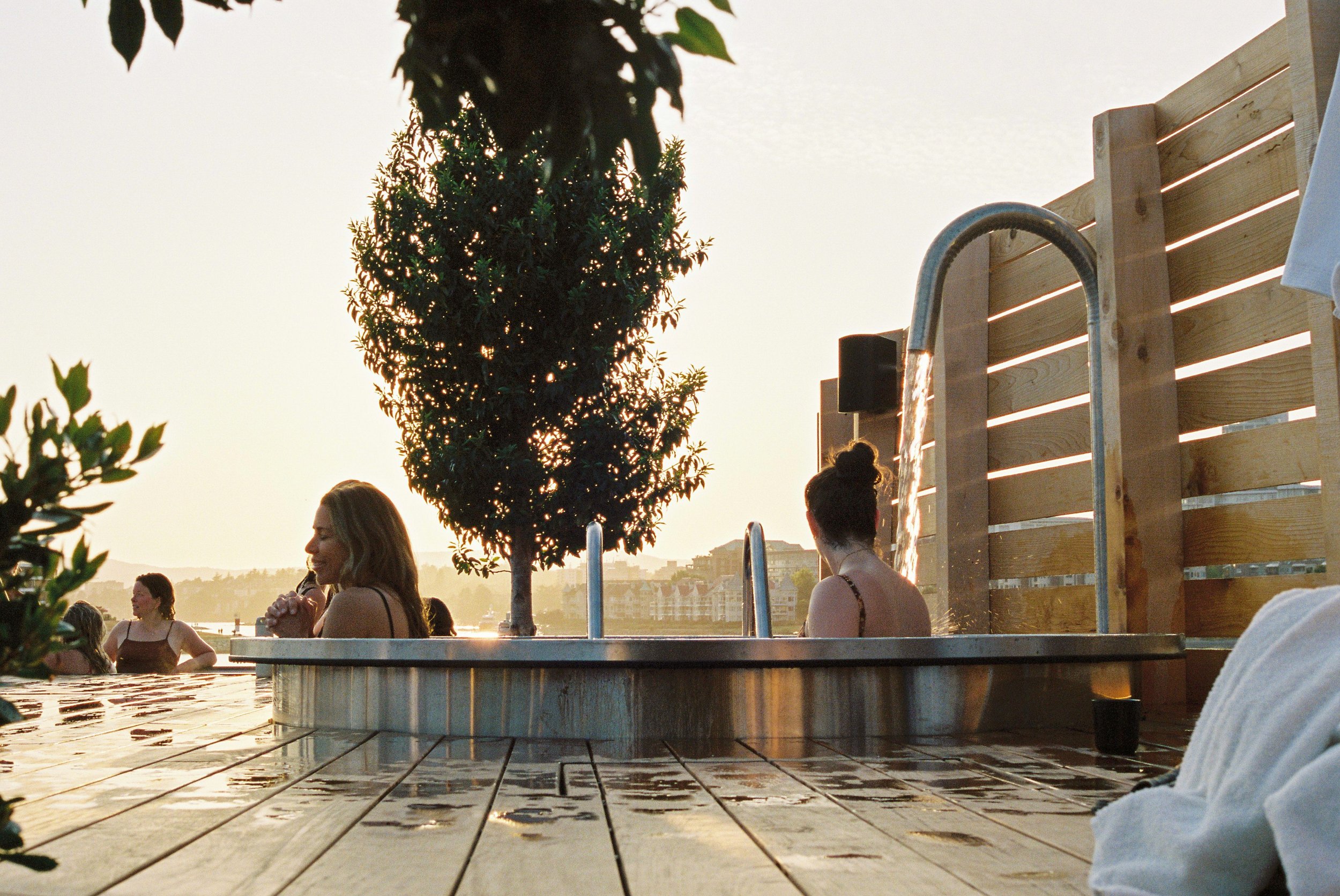 People relaxing in an outdoor hot tub with a wooden privacy screen, a tree, and a view of buildings across a body of water at sunset.