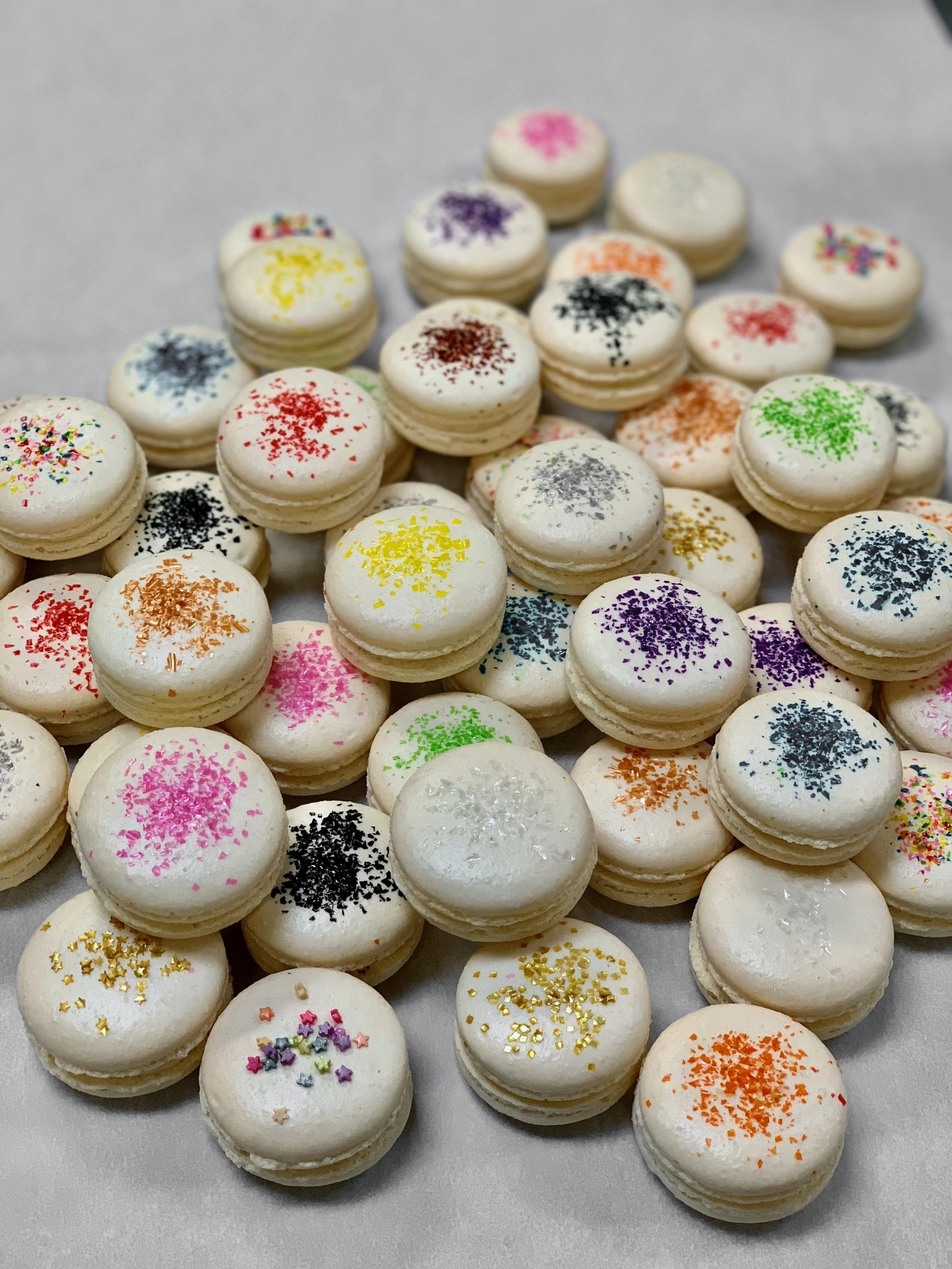 Assorted small macarons decorated with colorful sprinkles and glitter on a plain background.