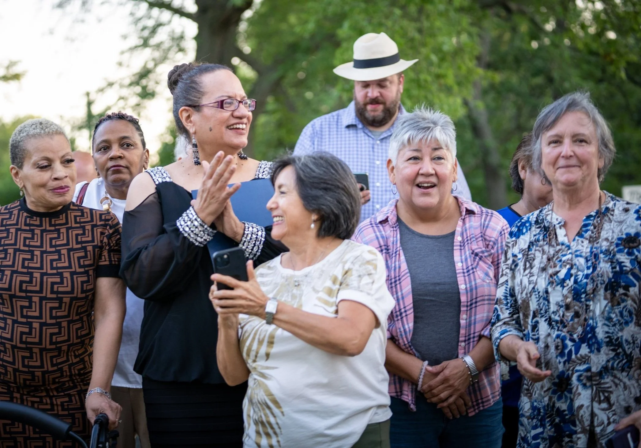 A group of diverse women and a man gather outdoors in a park-like setting, smiling and chatting during daytime.