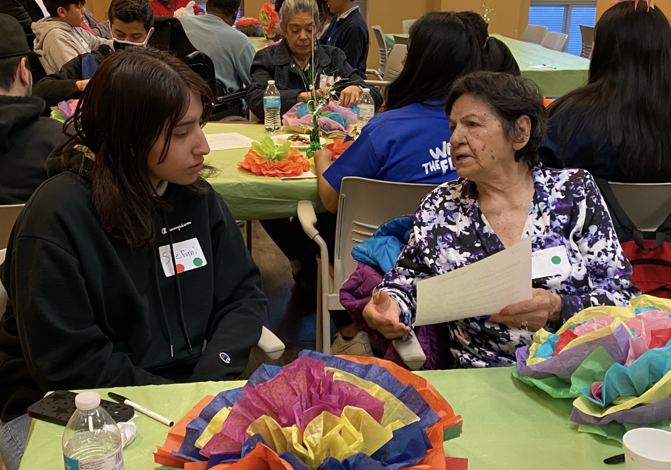 Older woman and young girl engaged in conversation at a colorful decorated community gathering, with multiple people seated at tables with vibrant tissue paper decorations.