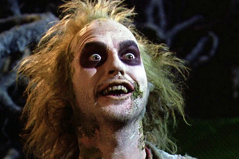 A character with wild, unkempt blonde hair, heavy dark eye makeup, and a wide smile showing uneven teeth.